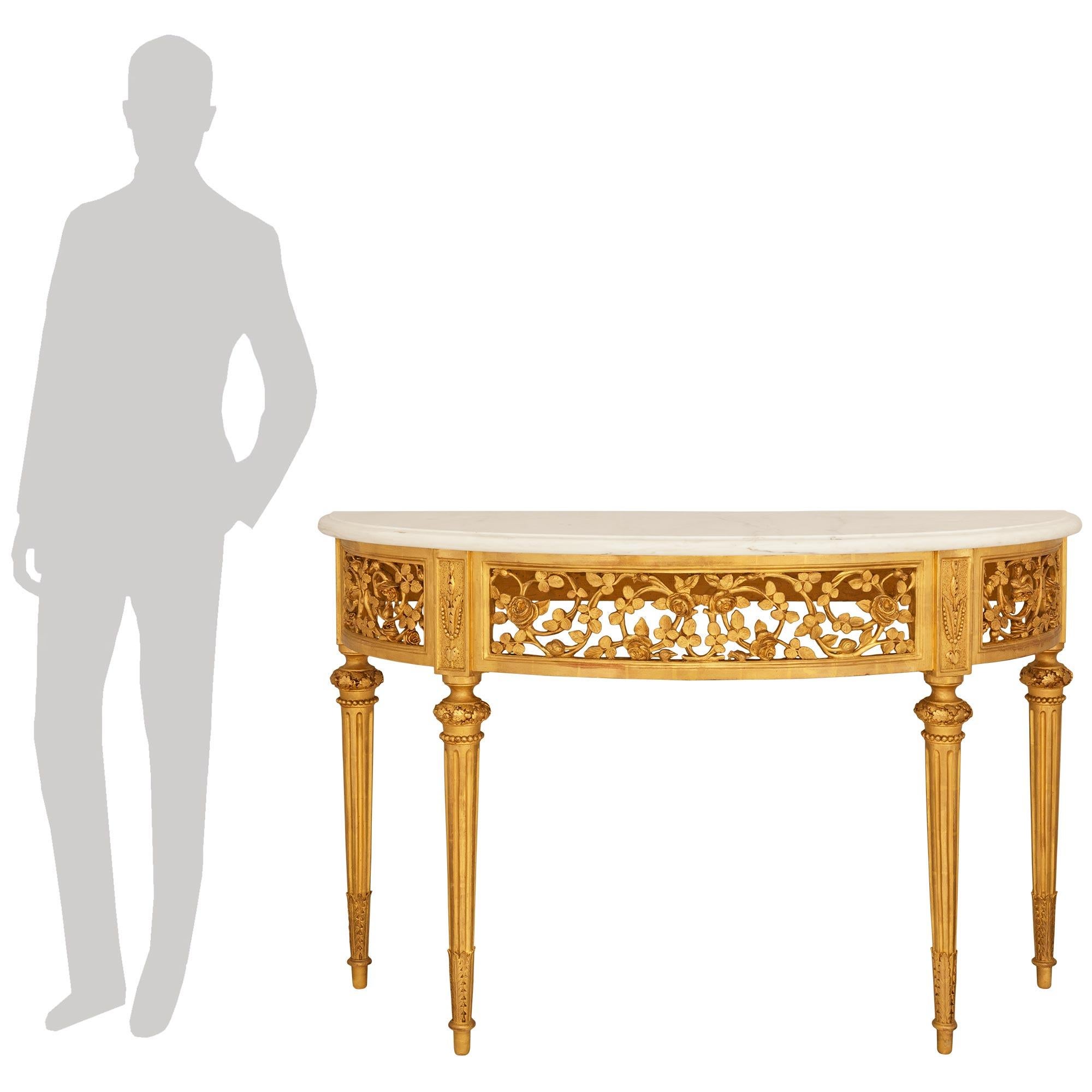 Espectacular y elegantísima consola francesa del siglo XIX, de estilo Luis XVI, de madera dorada y mármol Calacatta. La asombrosa consola en forma de demi luna se eleva sobre patas cilíndricas estriadas y ahusadas, con adornos de hojas de roble