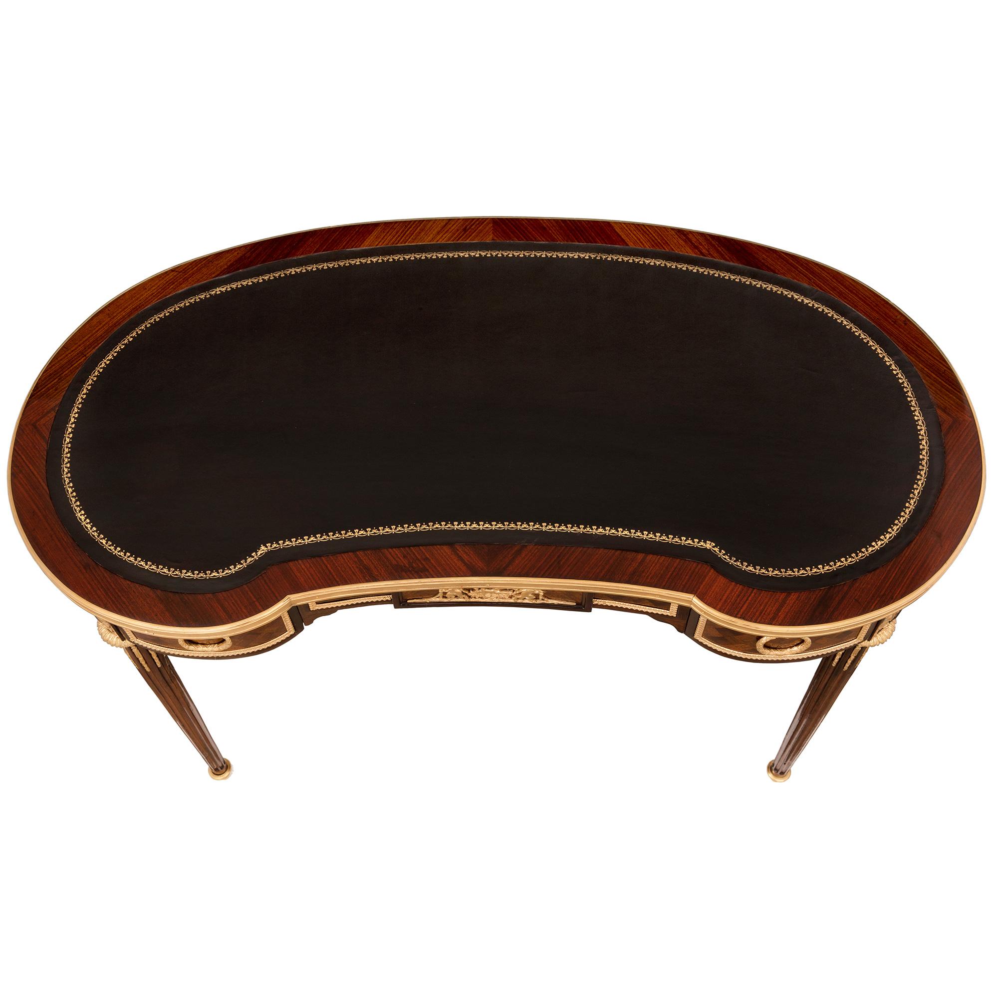 An elegant French 19th century Louis XVI st. mahogany, kingwood and ormolu kidney shaped writing desk. The desk is raised by slender circular tapered fluted legs with fine foliate topie shaped sabots, foliate top caps and richly chased fitted