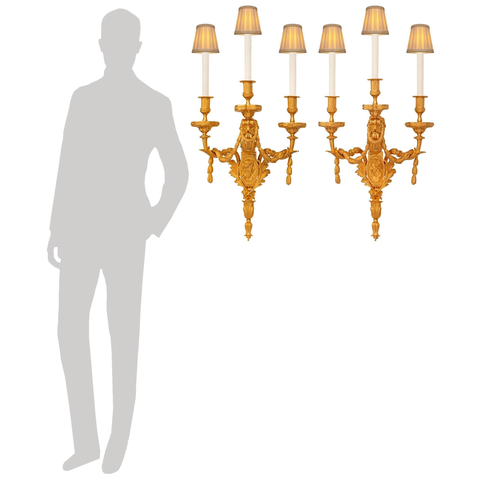 Pareja de apliques franceses de Ormolu del siglo XIX, de estilo Luis XVI, de gran tamaño y muy decorativos. Cada exquisito candelabro de tres brazos está centrado por remates inferiores decorados con follaje que se fijan a la fina placa trasera