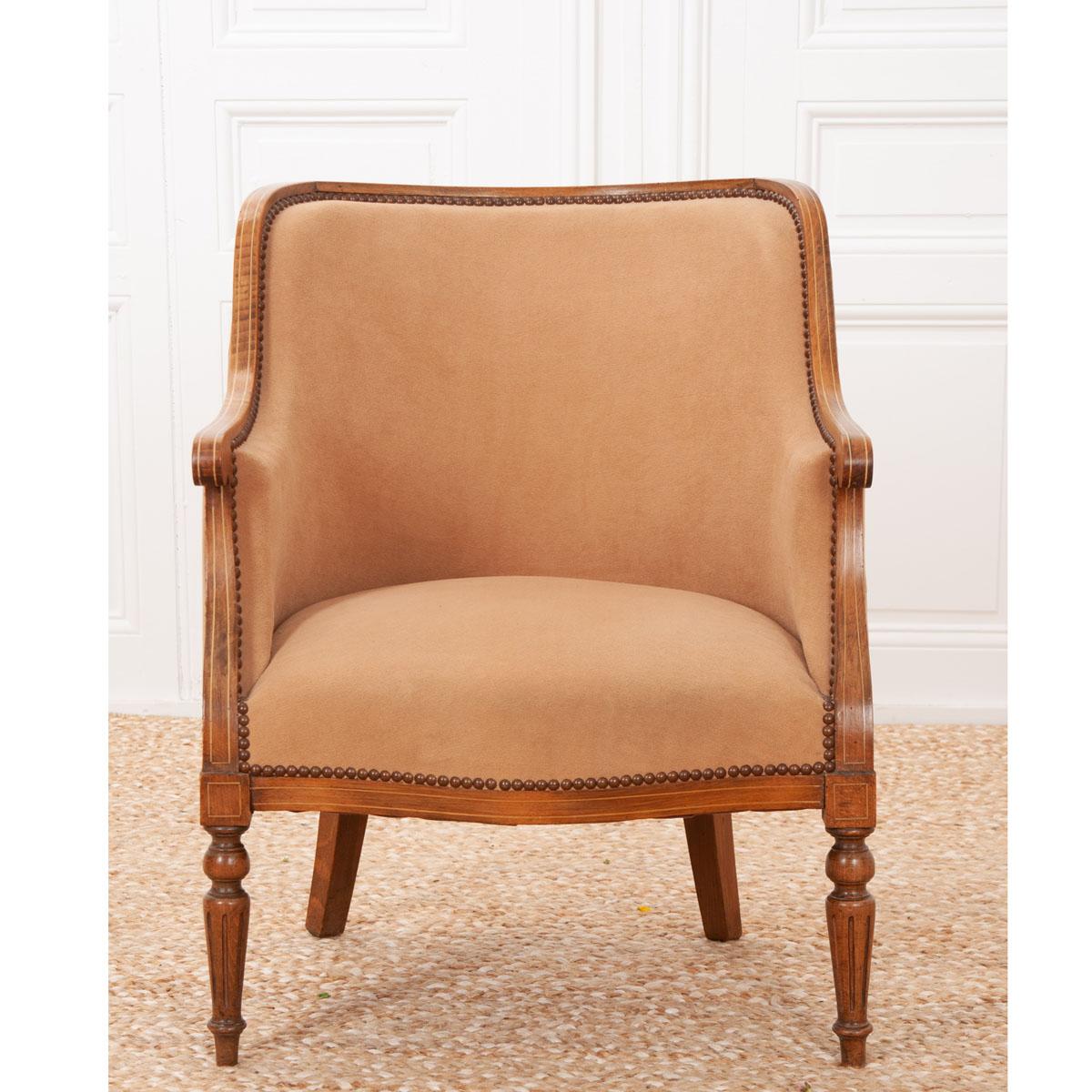 A single, inlaid walnut and upholstered Louis XVI-style armchair. The frame has a rich tone with a thin inlay throughout, wrapping around the back and down both arms. This chair is upholstered in a camel colored fabric that is trimmed in nail heads.