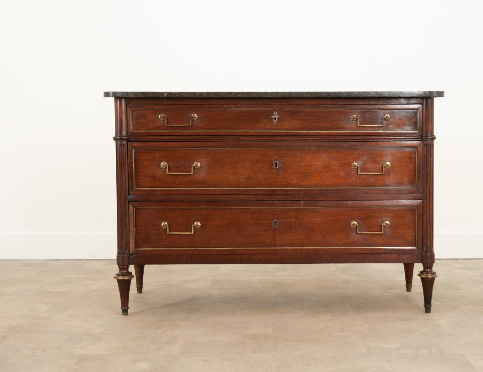 This Louis XVI style commode is made of mahogany, circa 1800, and topped with a shaped piece of Belgian Blue stone. A bank of three drawers with inset brass paneling are all fit with easy to use brass hardware. Each drawer features a functioning