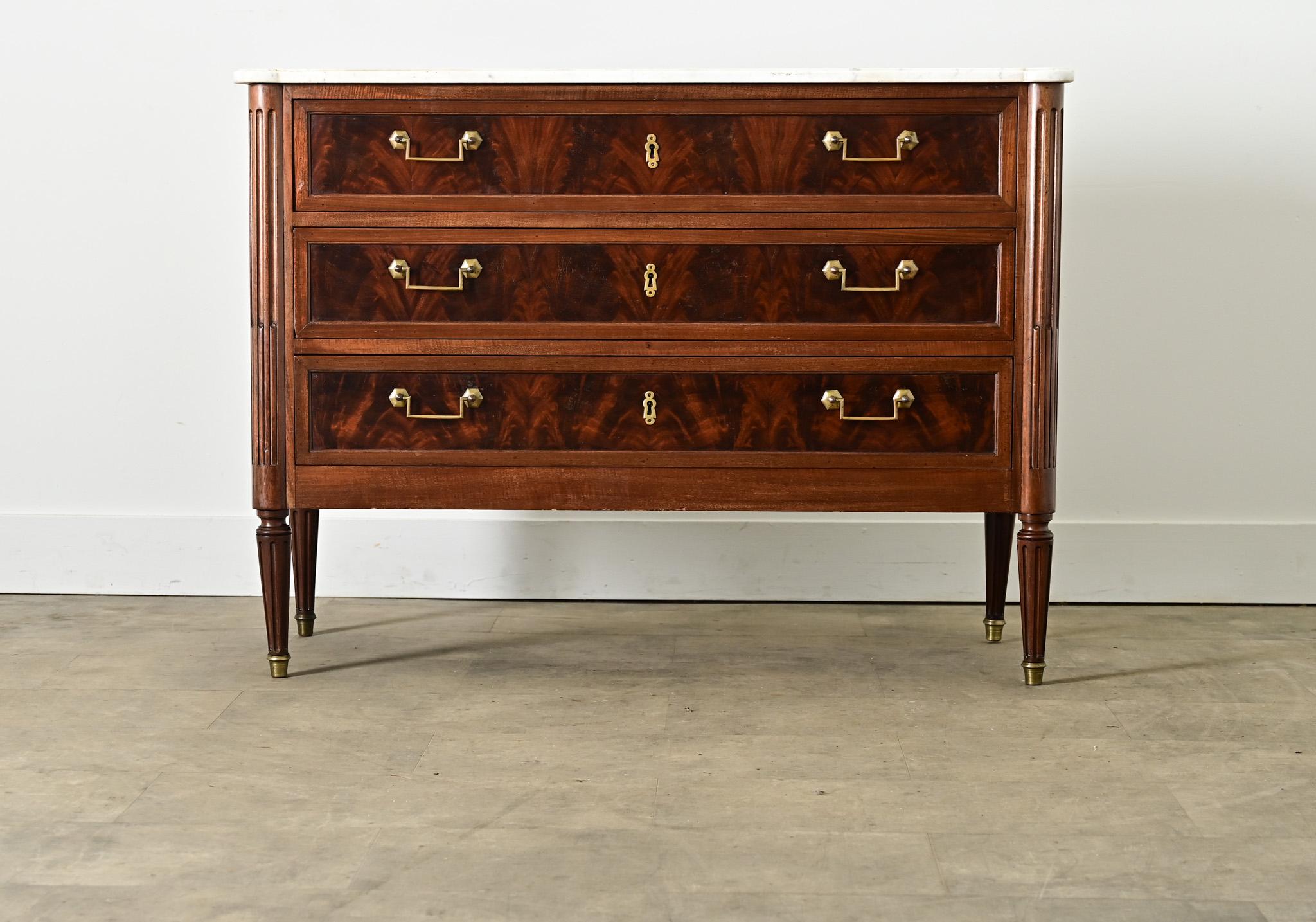 A magnificent 19th century Louis XVI-style commode from France, with a marble top on a mahogany case of three drawers. The white stone is traced with fine veining and a patinated surface of beige discoloration and scratches, carved in turreted front