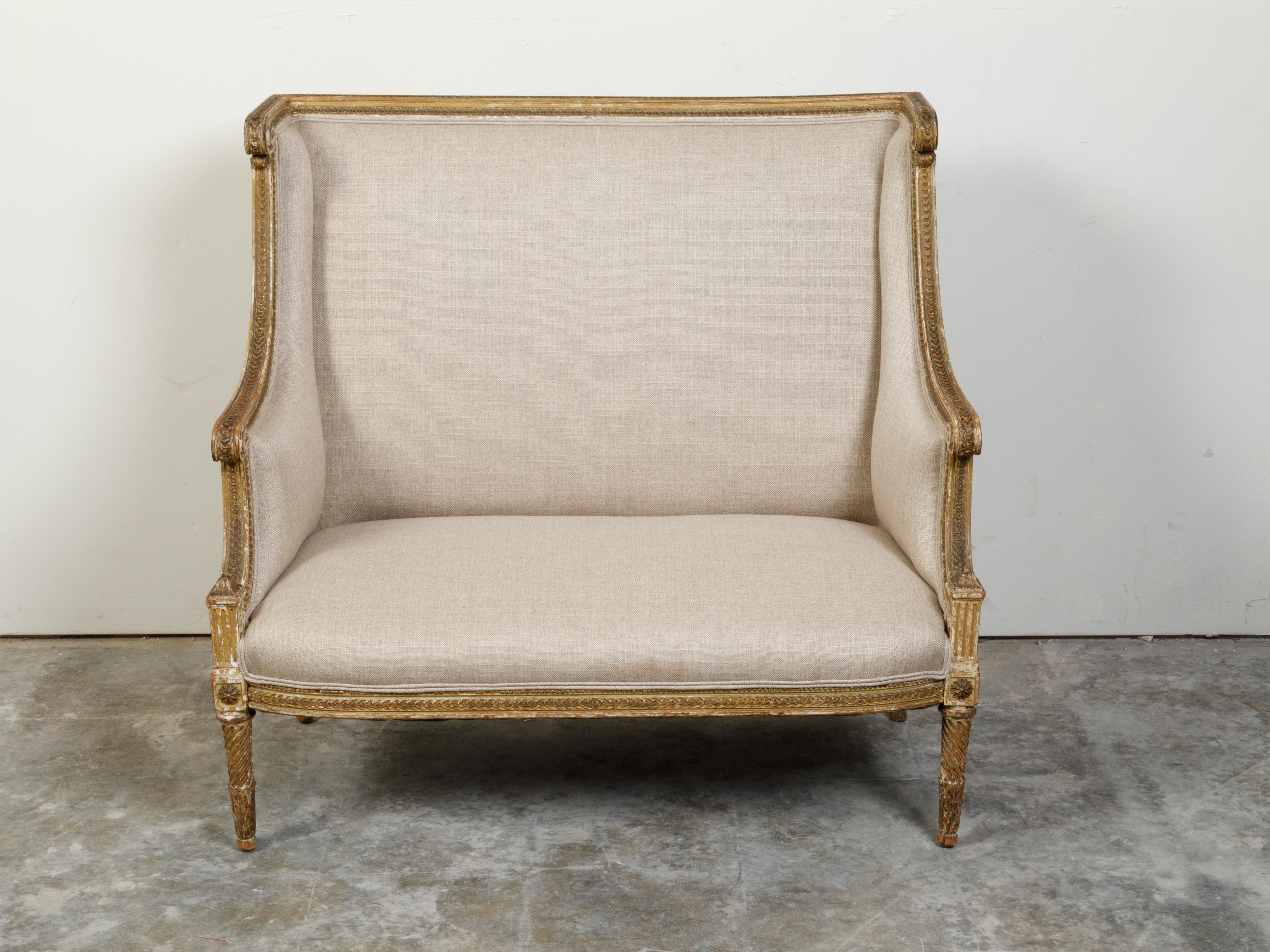 A French Louis XVI style giltwood wingback settee from the 19th century, with carved foliage motifs and distressed patina. Created in France during the 19th century, this giltwood settee features a wingback connected to scrolling arms flanking a