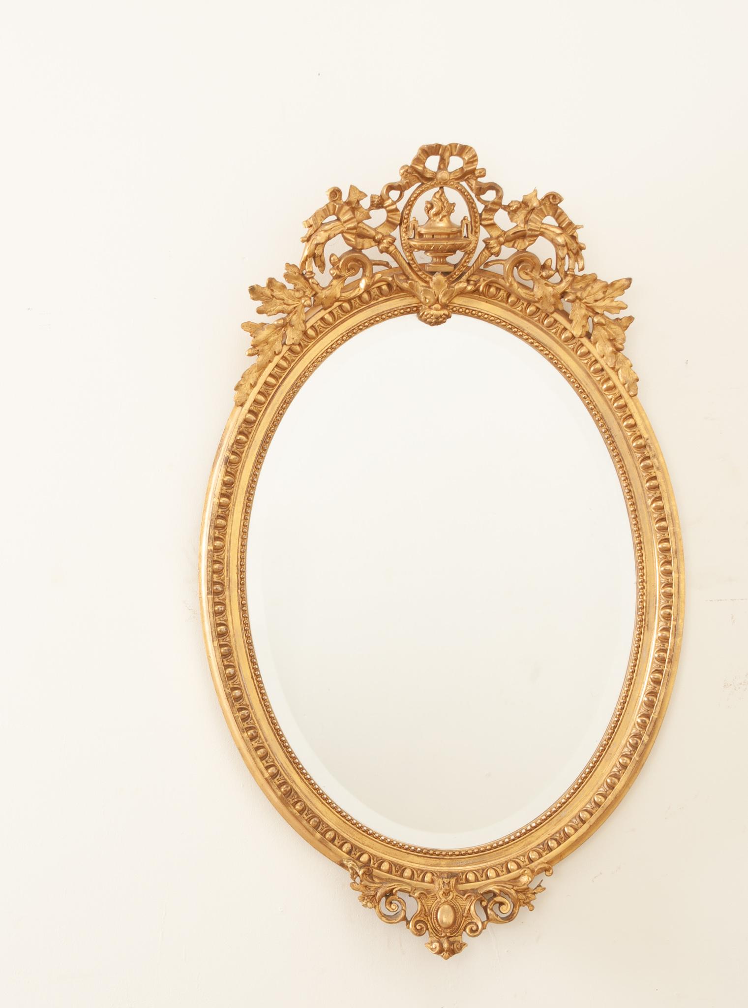 This beautiful antique gold gilt oval mirror was hand-crafted in France in the 19th century. A great example of Louis XVI style, the top ornament is a richly carved and out-sized asymmetric crest surmounted by a flambeaux-topped classical urn