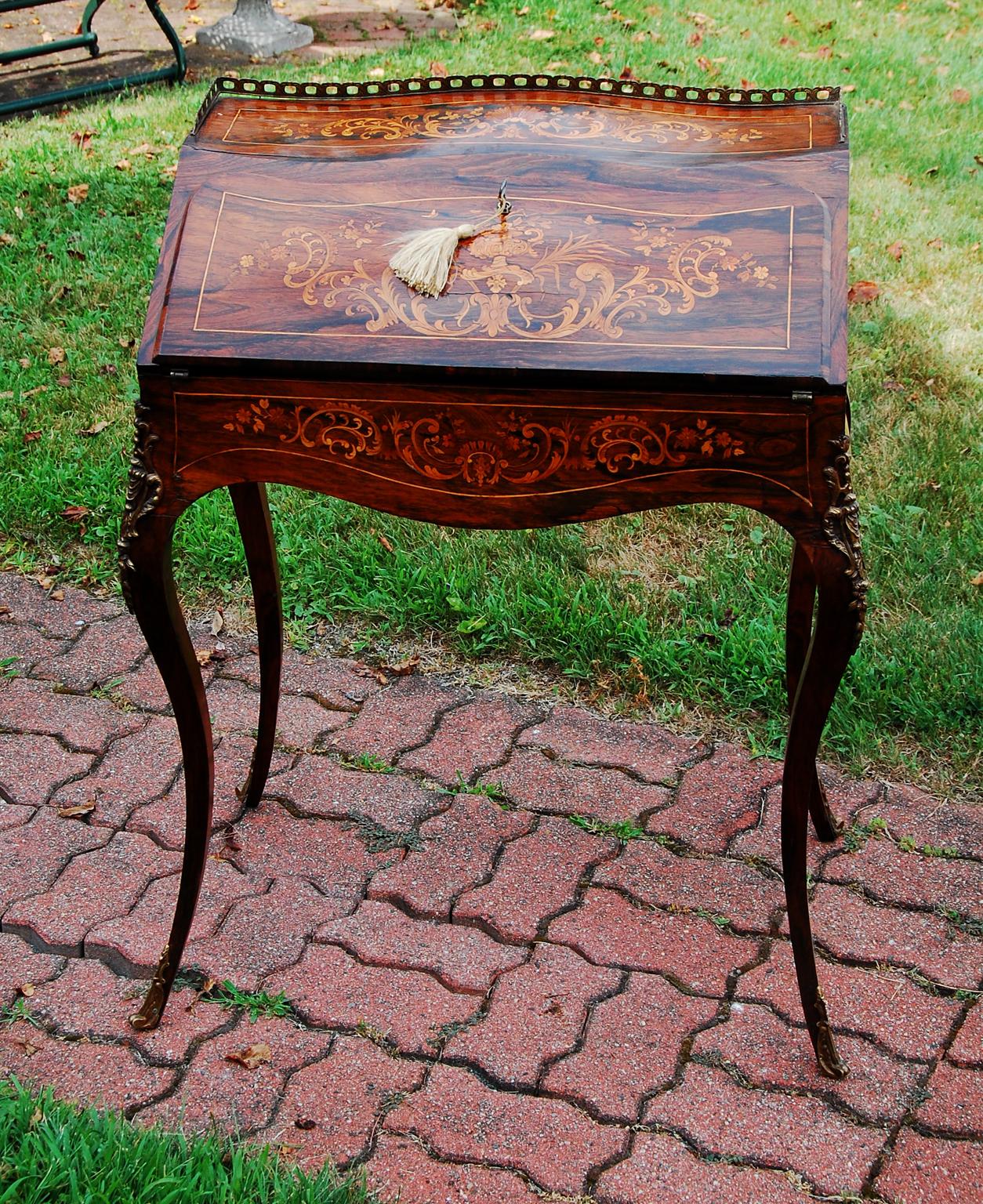 French Louis XVI style fall front serpentine lady's writing desk in rosewood with elaborate marquetry inlay in boxwood and satinwood. This diminutive, bombay, elegant writing desk has delicate cabriole legs with ormolu mounts and a brass gallery to