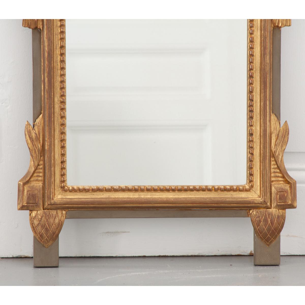French 19th century Louis XVI-style mirror has a gold gilt finish with accents of gray-green paint. It has a wood carved crest of an urn with a floral bouquet and swags hanging down on either side. This petite mirror is very stunning.
