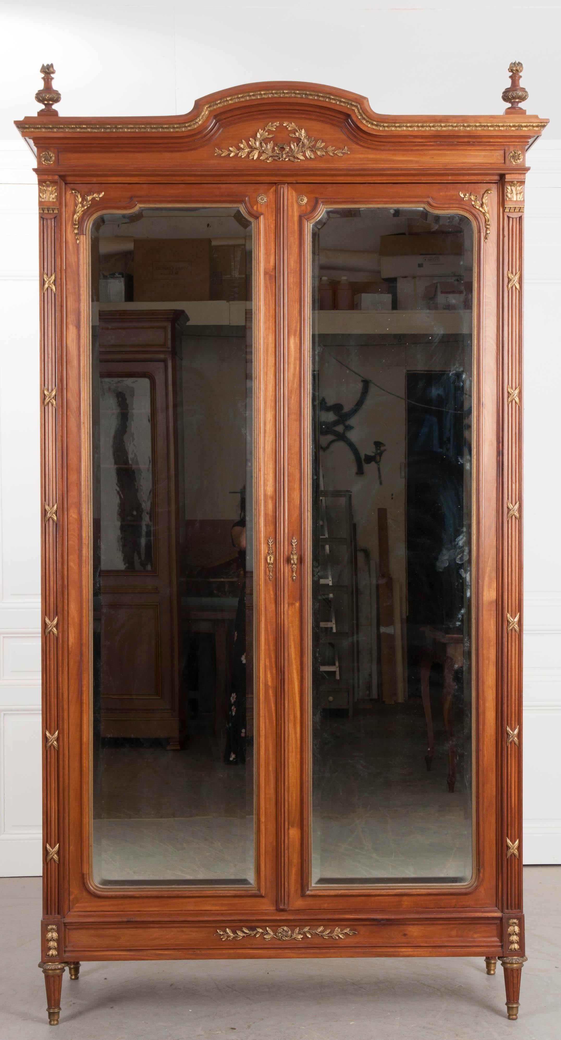 A handsome Louis XVI-style armoire, with mirrored doors, from 19th century France. The brass-trimmed and shaped cornice is bedecked with urn and flame finials on top, and a gilt brass ribboned wreath found in the frieze. The tall doors have shaped,