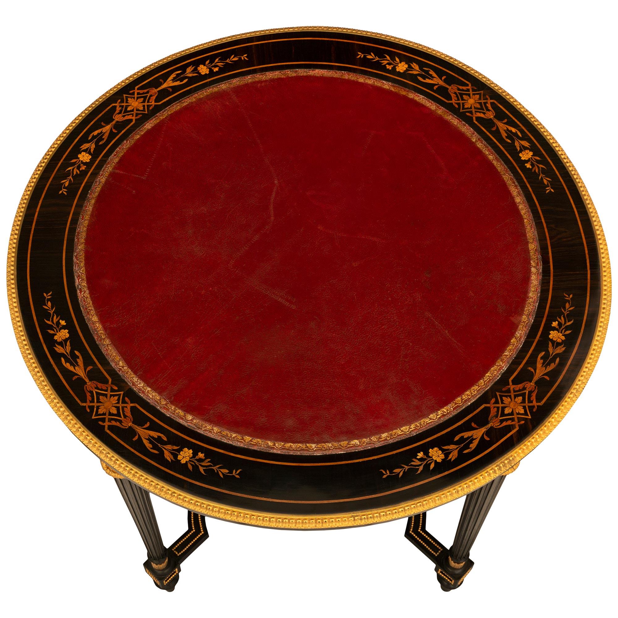 A striking French 19th century Louis XVI style Napoleon III period ebonized fruitwood, exotic woods, ormolu and leather center table. The table is raised by elegant circular fluted legs with foliate top caps and fine toupie shaped feet. Each leg