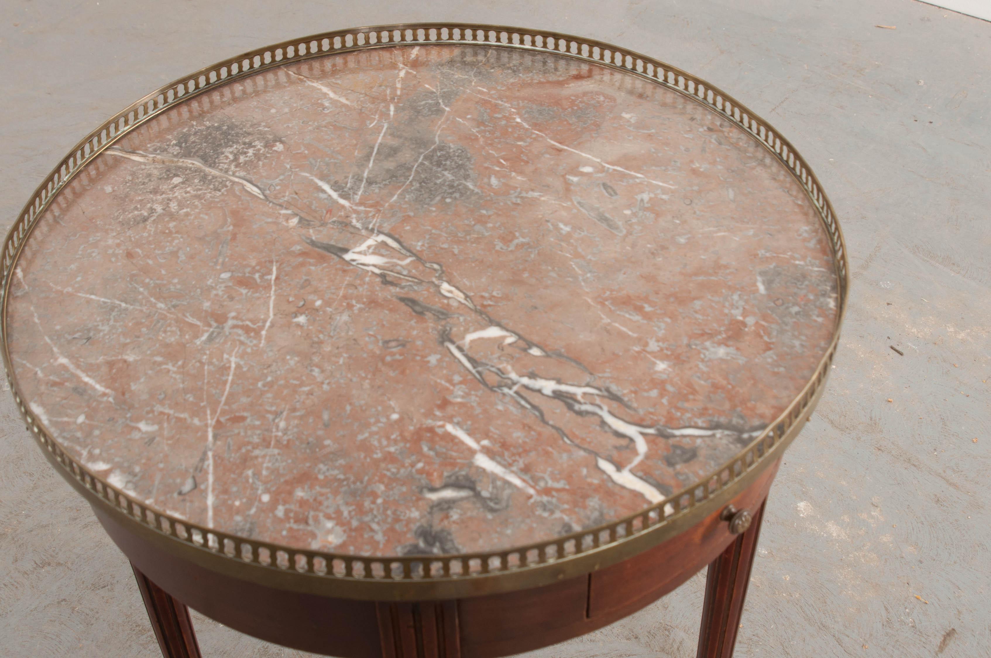 This fine Louis XVI-style circular mahogany gueridon table, circa 1880, is from France and features a gorgeous rouge marble top with grey and white veining, and reticulated brass gallery, above a pair of opposing apron drawers, raised on fluted and