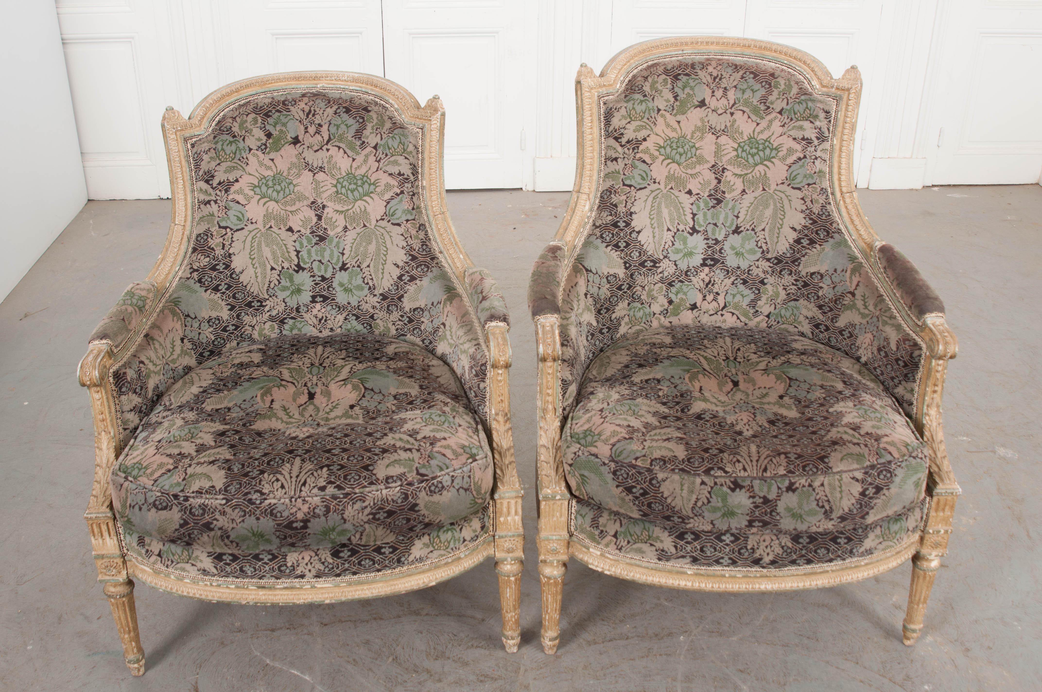 This elegant pair of Louis XVI-style cream-painted “His” and “Hers” bergères, circa 1860s, are from France and feature gorgeous velvet floral upholstery in shades of aubergine, lavender, grey, and green. The painted frames are highlighted in a sage