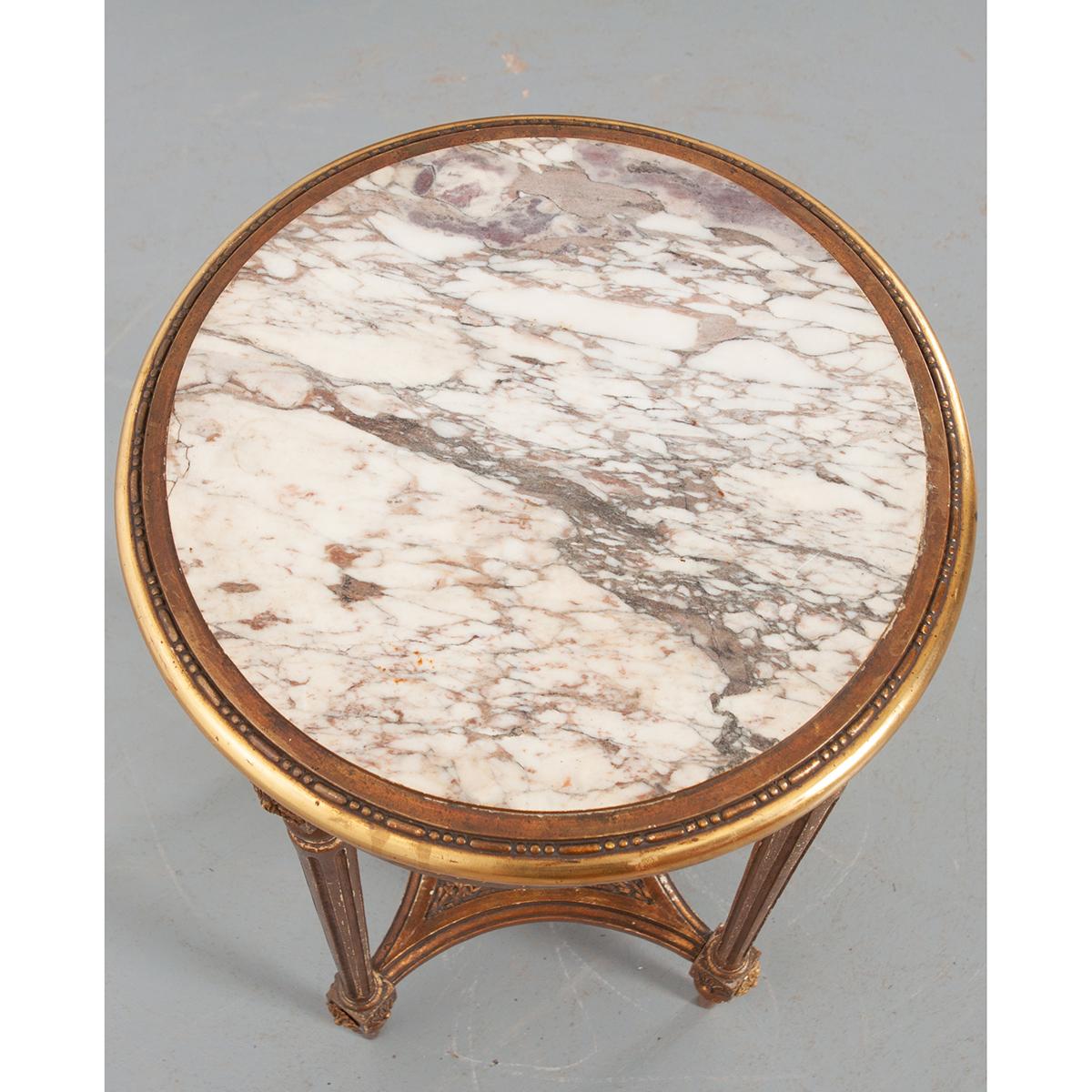 This elegant Louis XVI style gold gilt side table features a beautiful marble top over a ribbon-carved apron, supported by turned, tapered and fluted legs with rosette dies at each end. The four legs are joined by a four sided, concave shape caned