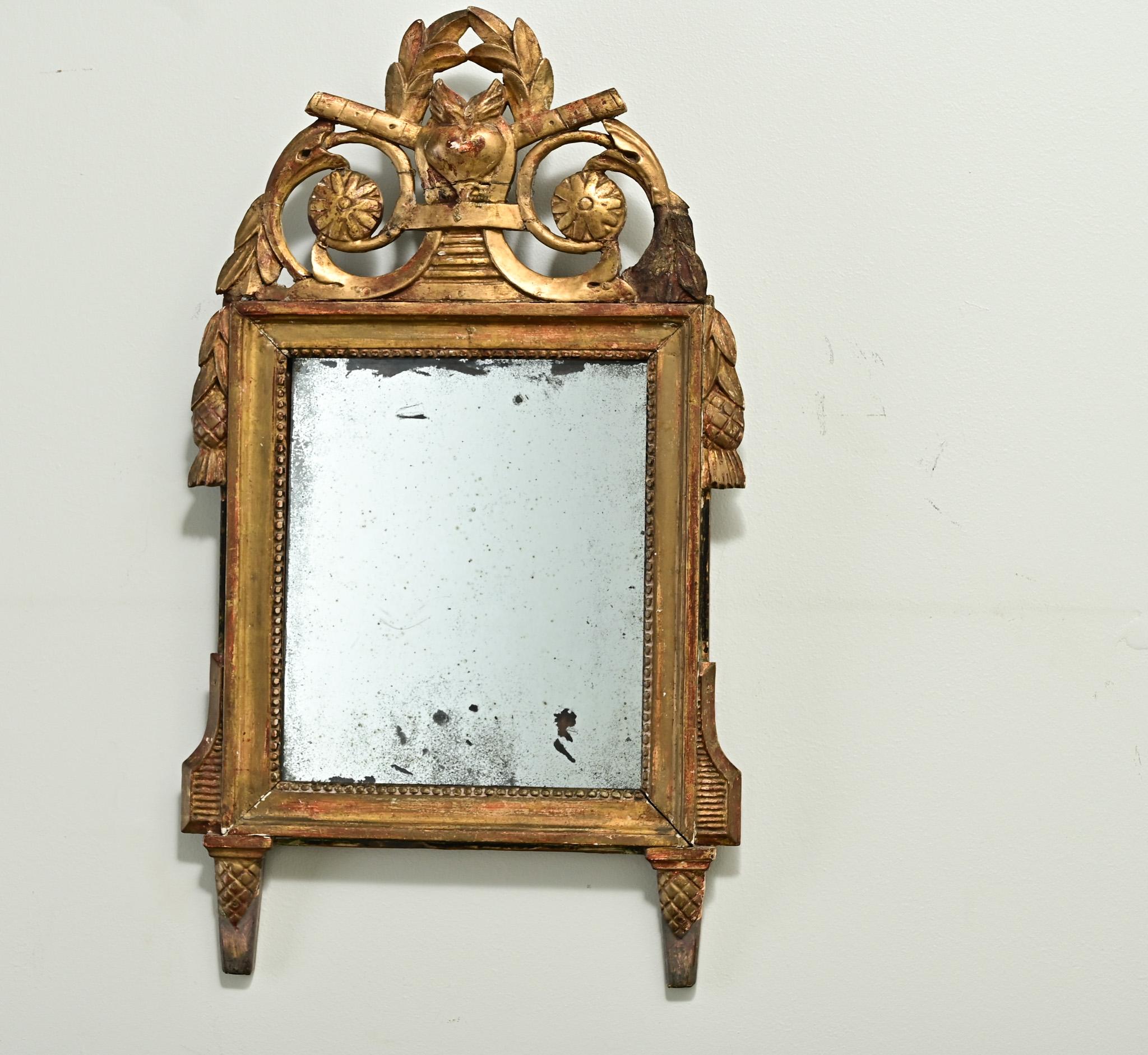Un piccolo trumeau francese in stile Luigi XVI, dorato. La cornice dello specchio, dorata e dipinta, presenta uno stemma intagliato con un cuore fiammeggiante e foglie di alloro. La lastra a specchio originale presenta foxing, scintille ed è
