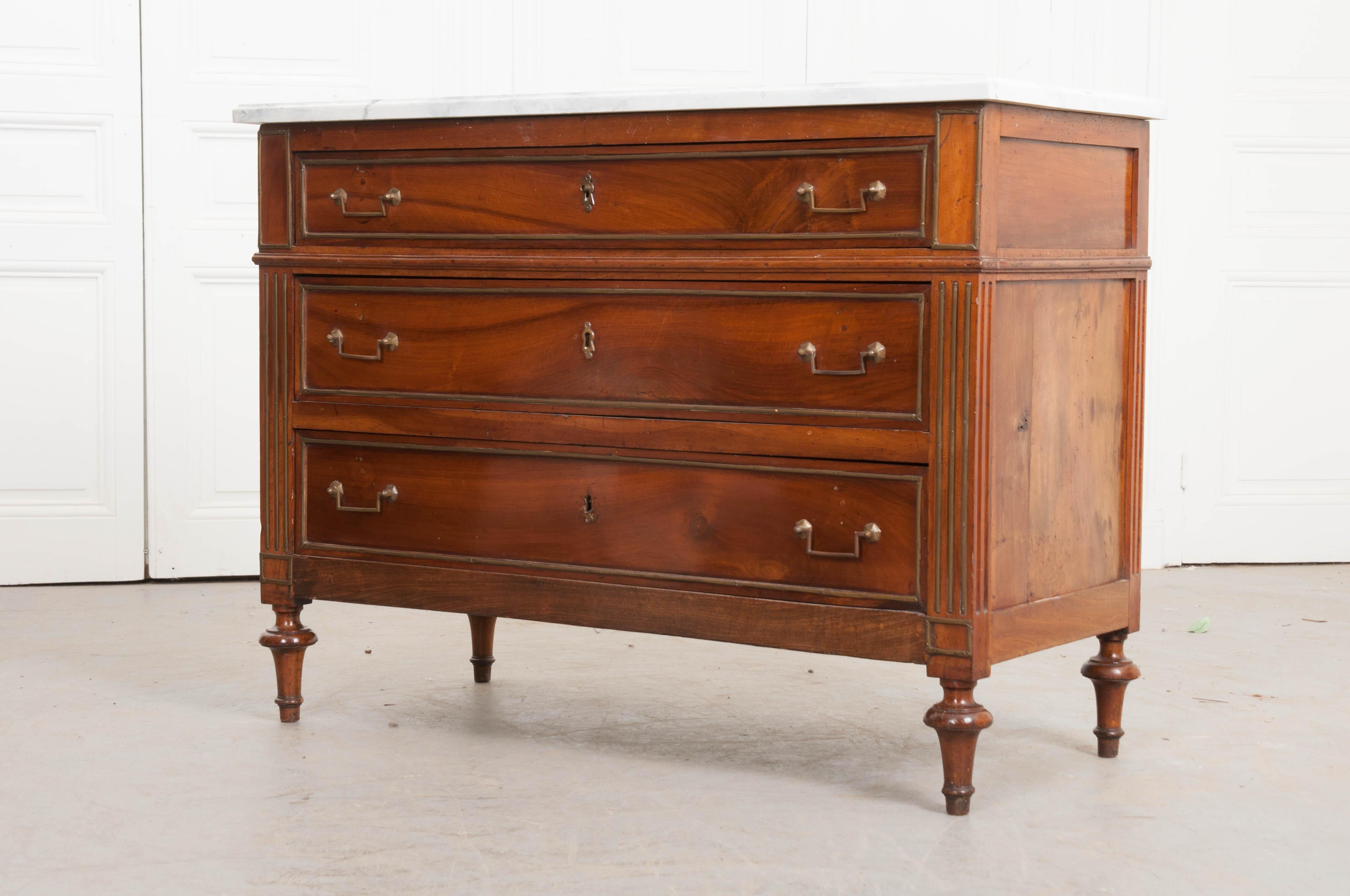 This Louis XVI-style walnut commode with white faux marble top, circa 1890, is from France. It features three drawers, the fronts of which are trimmed with brass and have brass-pull handles and escutcheons, flanked by fluted pilasters and raised on
