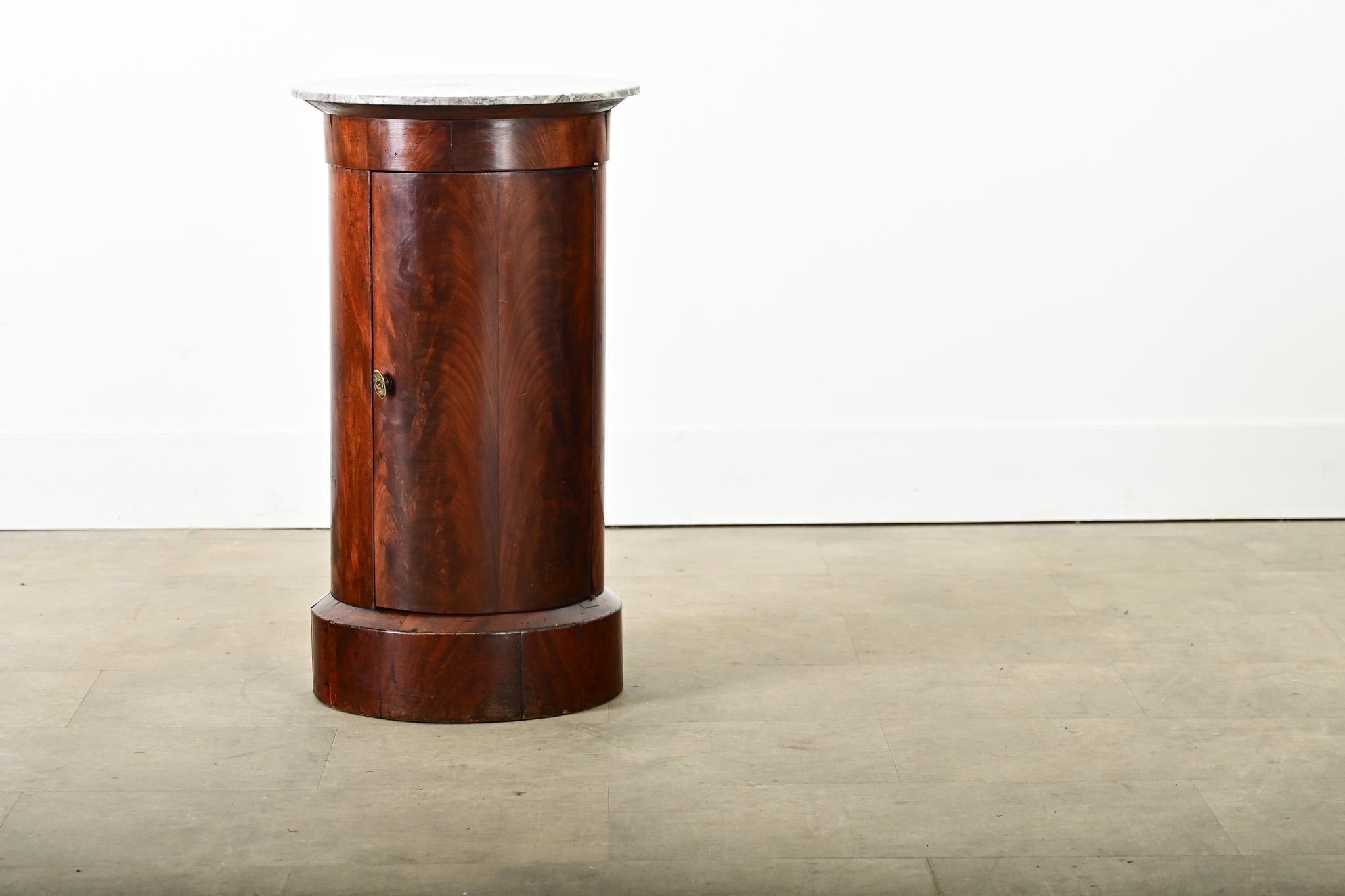 A columnar, French mahogany and marble top bedside table that offers both storage and display to fit in any room around the home. The top surface is a flat disc of pale gray marble traced with dark gray veining. Inside the exotic-looking mahogany