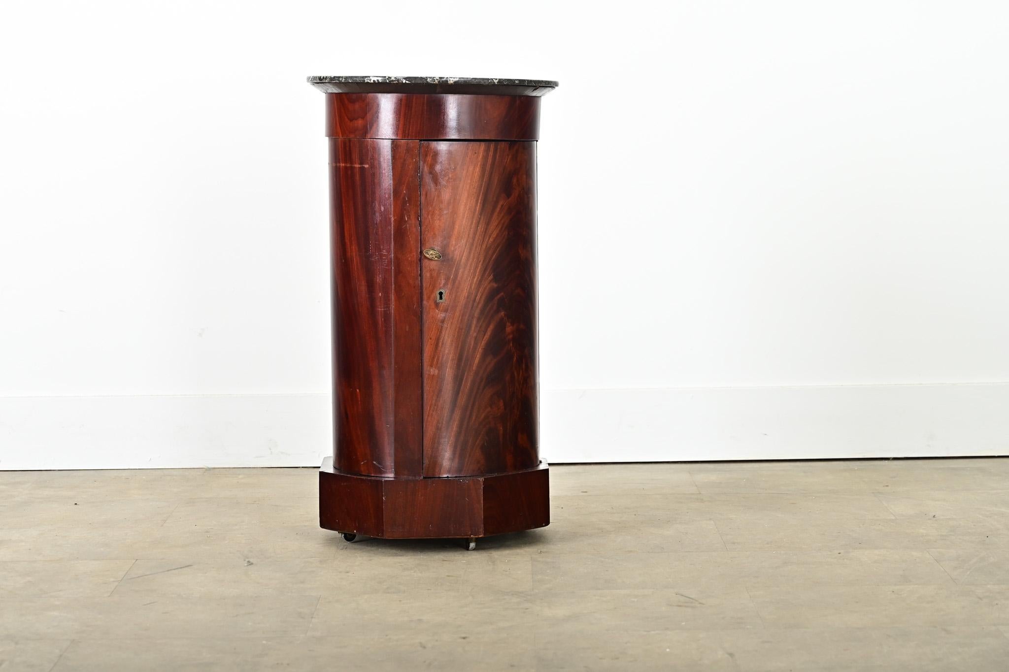 A columnar, French mahogany and marble top bedside table that offers both storage and display to fit in any room around the home. The top surface is a flat disc of Saint Anne marble, etched with scratches and chips after nearly two centuries of use.