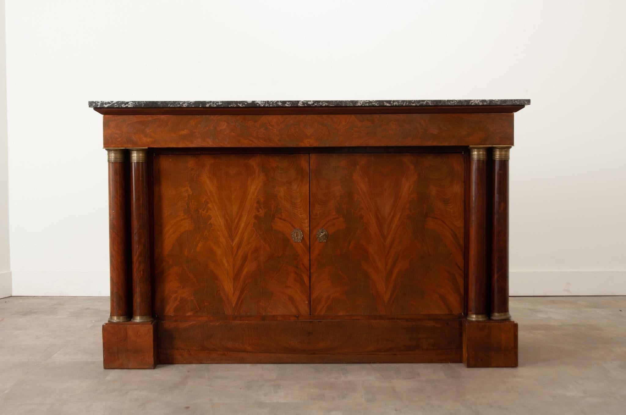 A French Empire buffet made of rich flame bookmatched mahogany is classic in style. The original marble top of charcoal with white veining sits at the top of this buffet and is removable. Below is a single wide drawer decorated with a burl