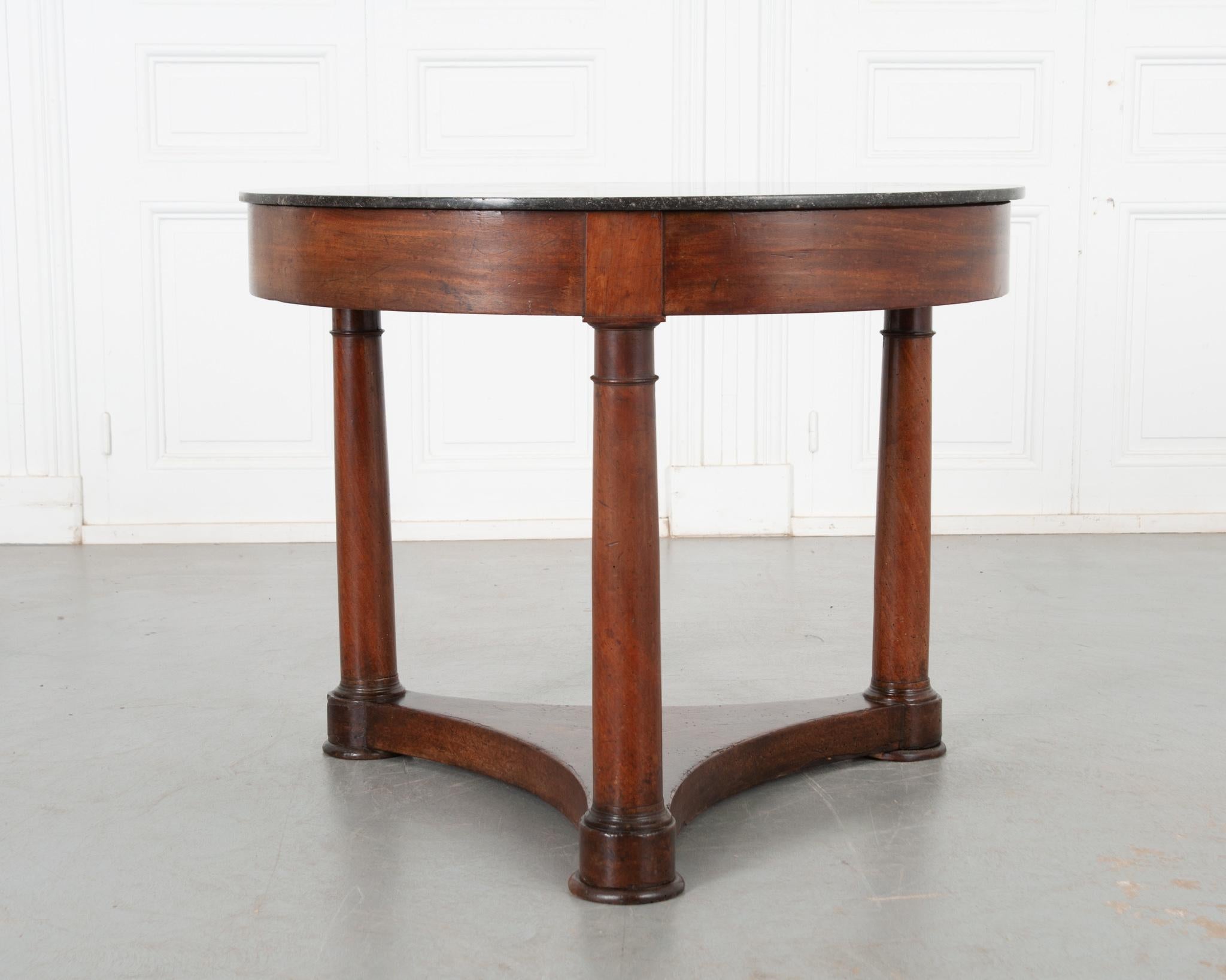 A sizable Empire style gueridon table from 19th century France. Beautiful black fossil marble top over a patinated mahogany body. A simple apron showcases the deep toned wood. Three columnar legs connect to a concave shaped stretcher that sits flush