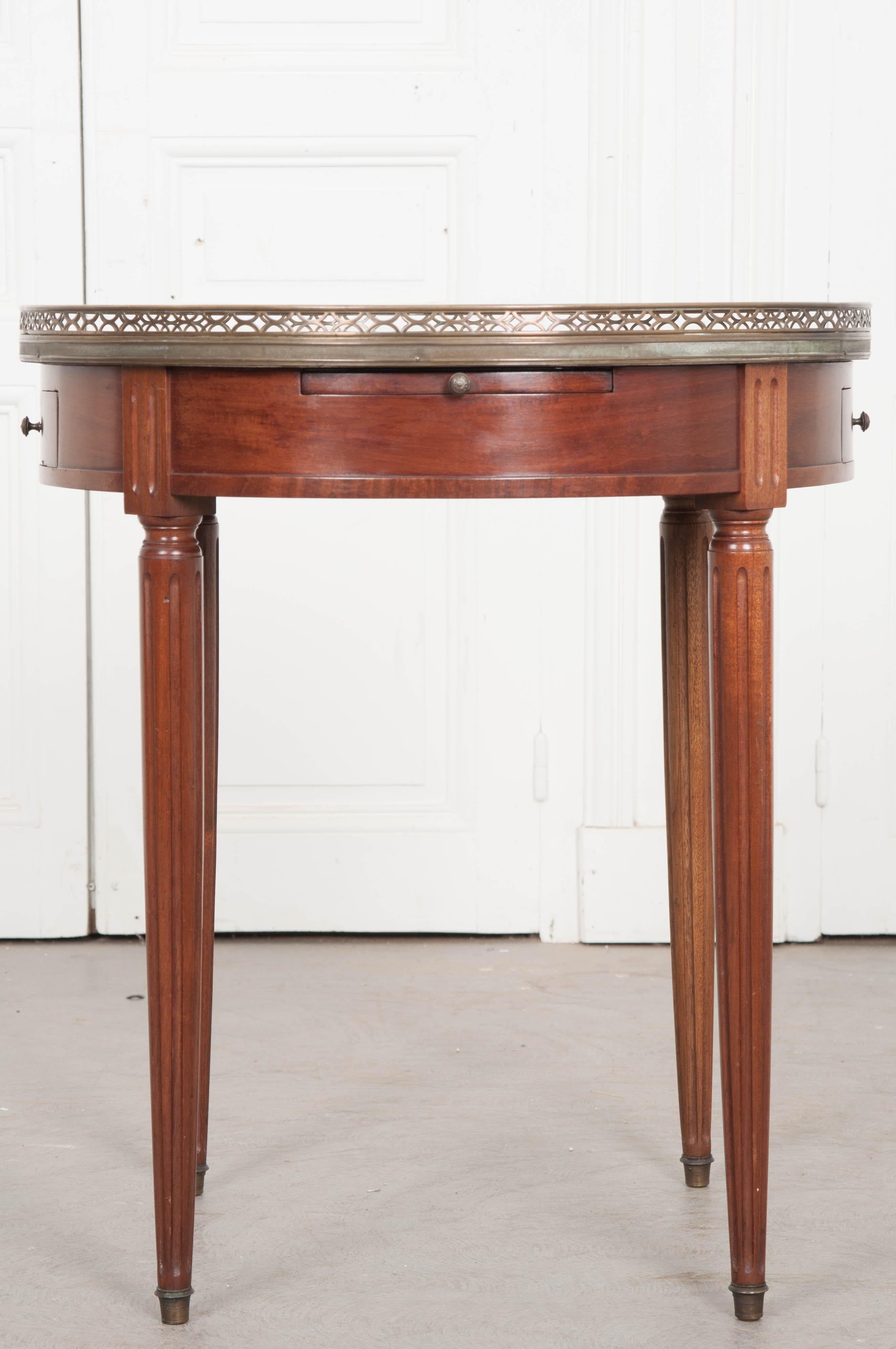 A classic example of a Louis XVI style marble-top guéridon, or circular side table, with brass gallery, is from France, circa 1880. The gray marble top with reticulated brass gallery rests over an apron having two drawers and two leather-topped