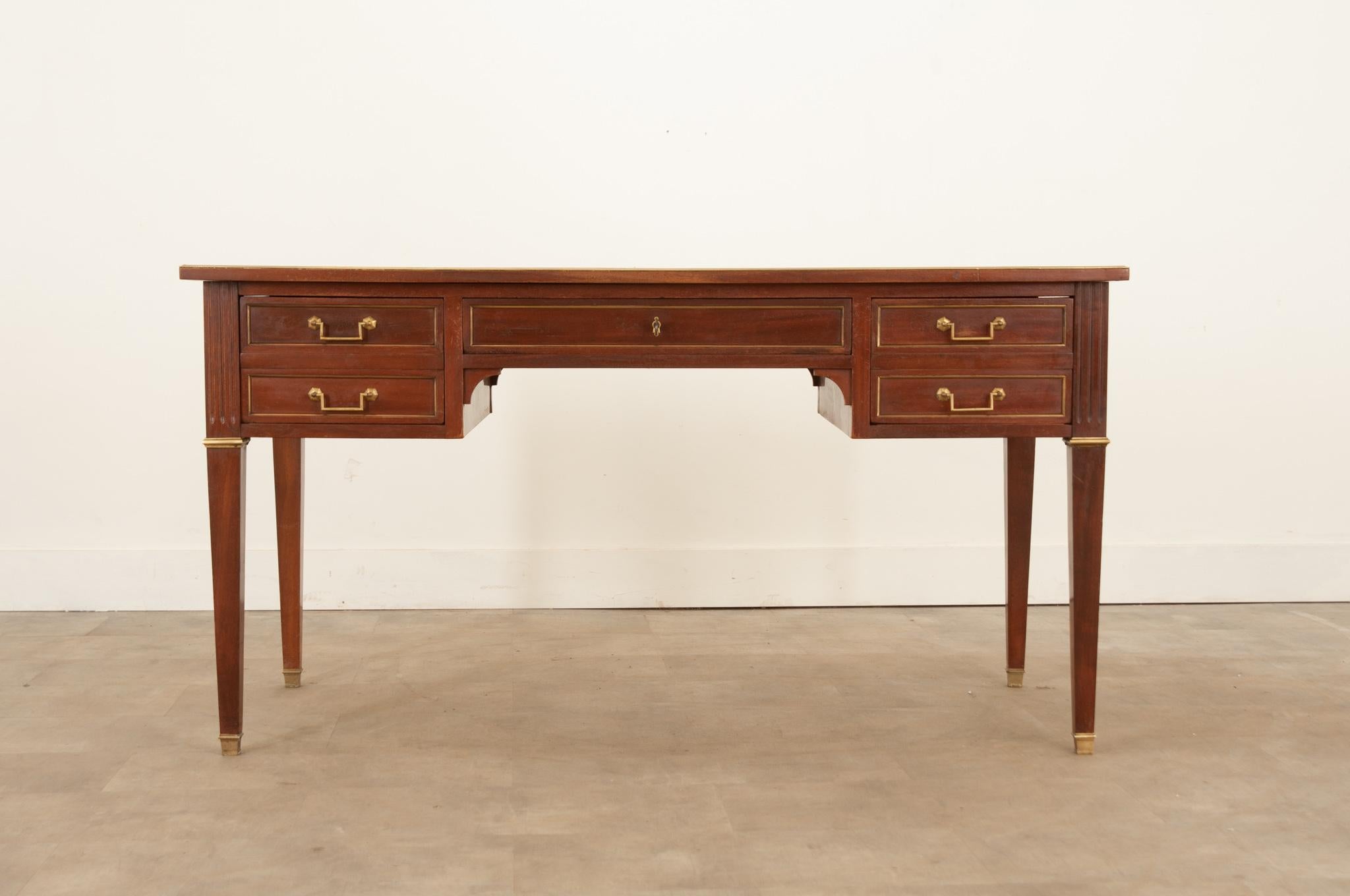A smart mahogany desk - with leather writing surface - from 19th Century France. This beautiful antique has a unique, transitional design that is heavily influenced by Louis XVI style. The top and drawer fronts are all trimmed in brass, and the legs