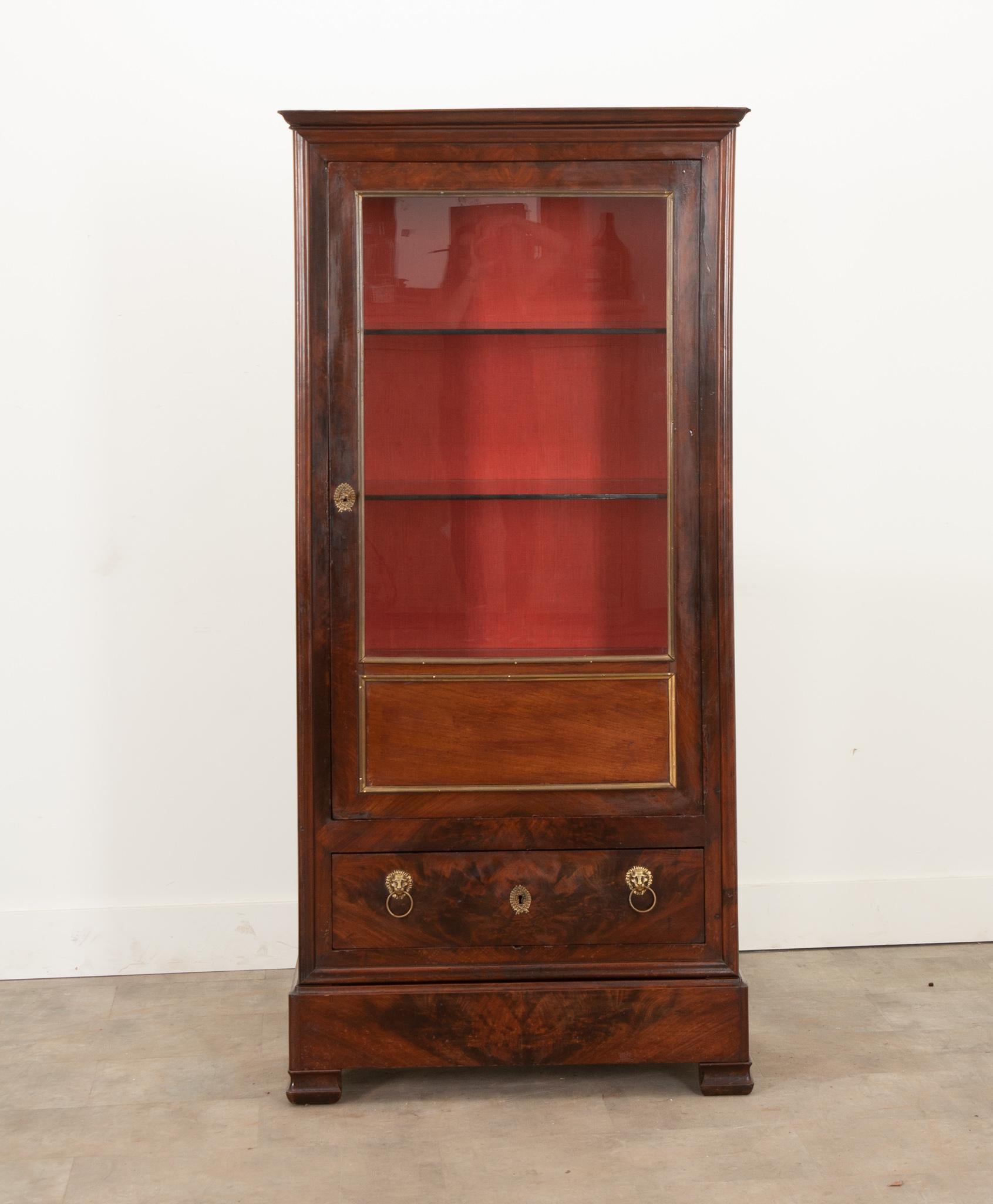 This Louis Philippe vitrine was crafted in France during the 19th century from finley bookmatched mahogany. A brass border encompasses the large single pane of glass and faux panel below it. The interior is lined with a vibrant red linen blend