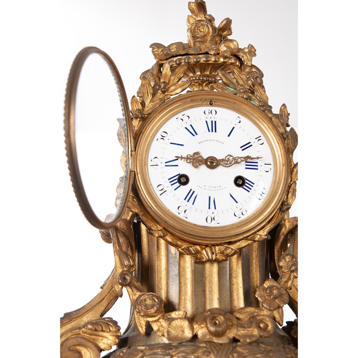 Französische Manteluhr aus Metall mit dem Aufdruck 'Boiscontier a Paris Rue De Caumartin' auf dem Zifferblatt. Das Wappen ist ein Korb mit kunstvollem Blattmotiv über einem symmetrisch geformten Gehäuse. Der Schlüssel betätigt das Uhrwerk der Uhr
