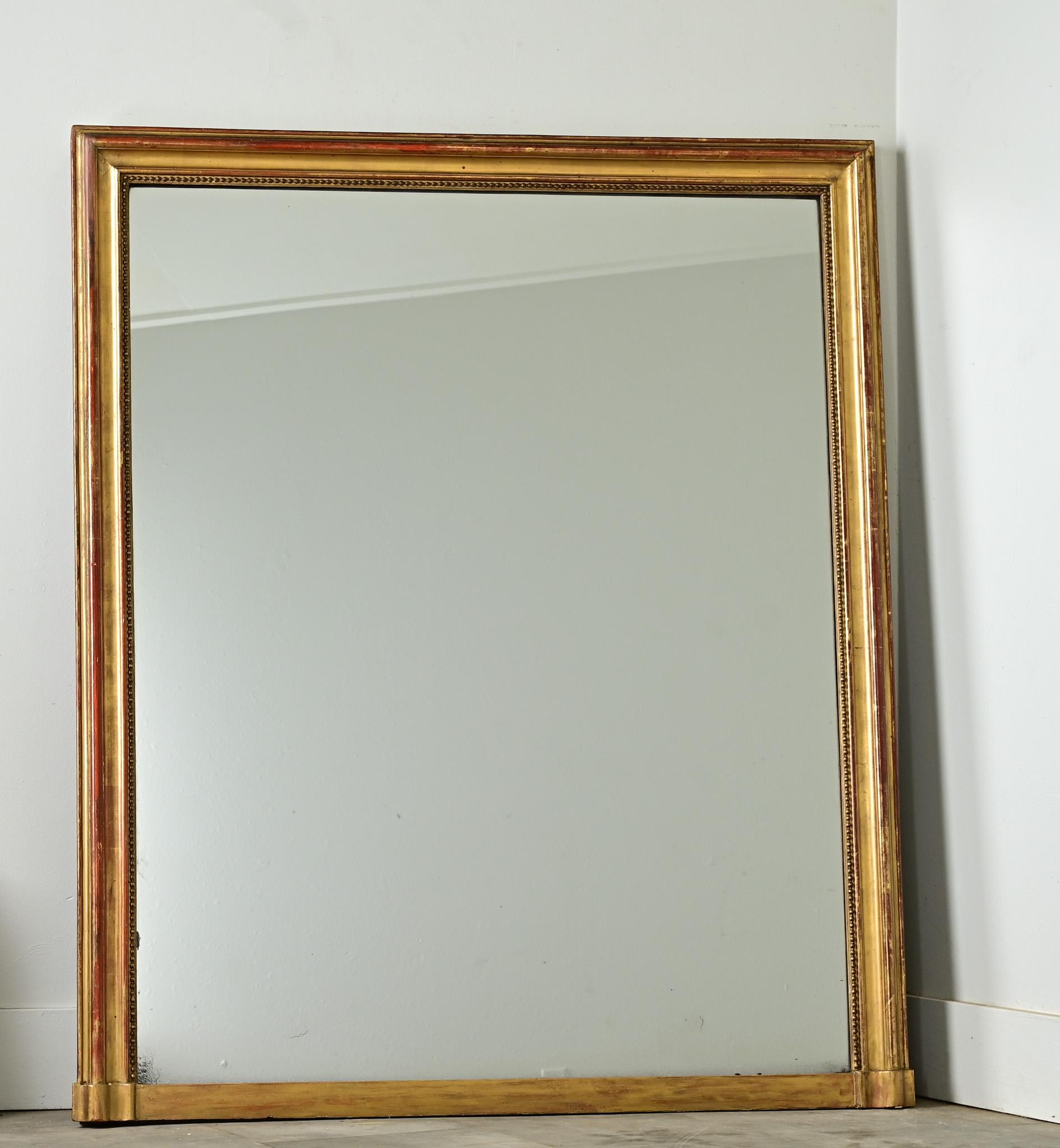 Strong proportions characterize this French mantel mirror, its large mirror plate framed by a classic and unassuming molding of velvety gold-gilt. Reflecting a mature patina, the frame blooms with red bole in a painterly touch. Foxing and