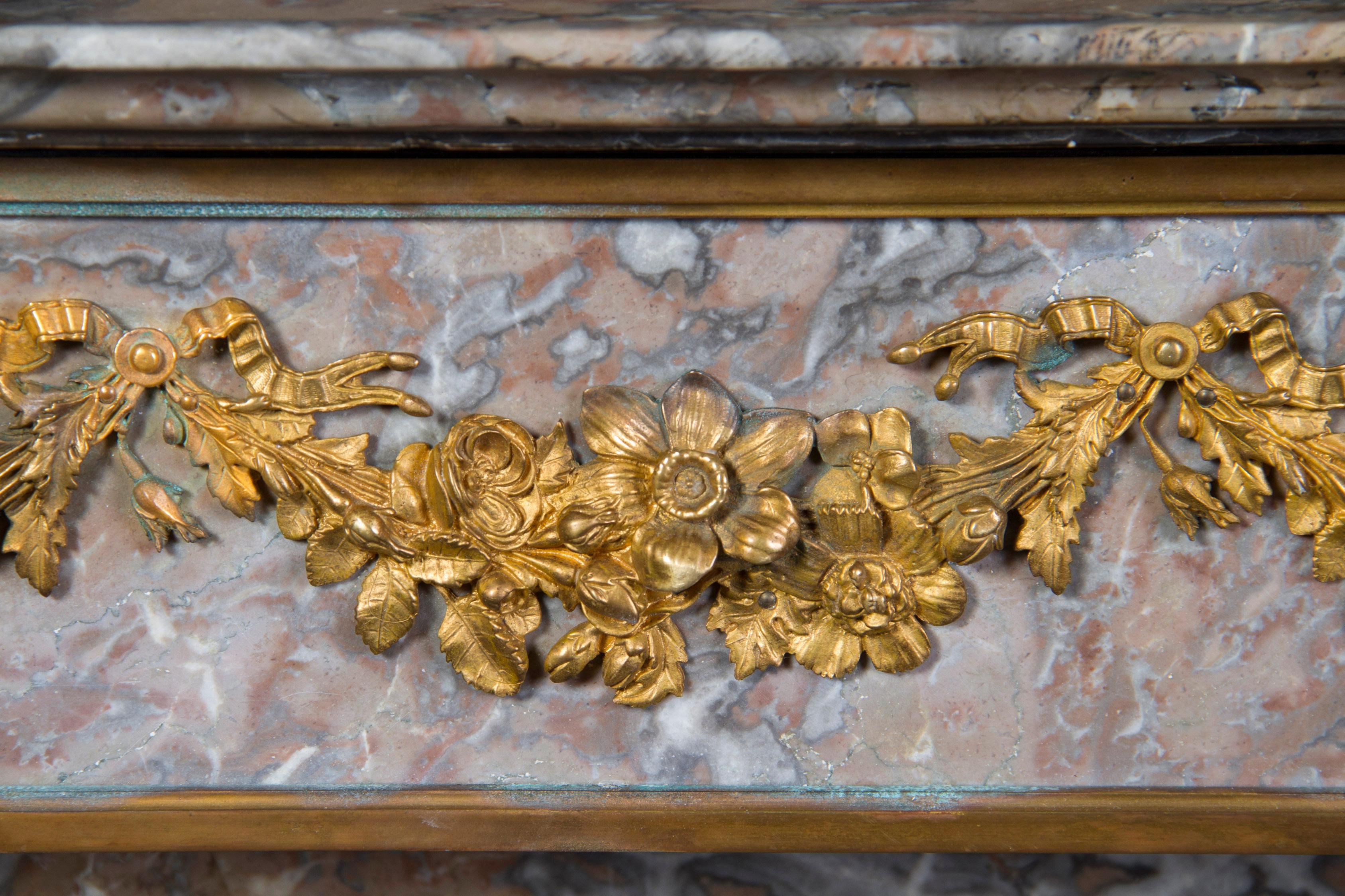 Questo bellissimo caminetto francese del XIX secolo Luigi XVI è caratterizzato da decorazioni in bronzo dorato con nastri e un disegno floreale. Le montature contrastano bene con le tonalità chiare di grigio e rosso del marmo. La curvatura del pezzo