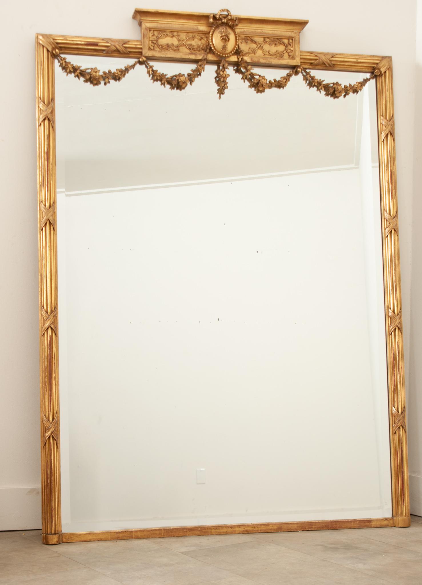 A gorgeous and regal Louis XVI style gold gilt mirror hand-crafted in France in the 19th century. This elegant mirror boasts the original mirror glass with a beautiful beveled edge. The hand-carved frame is topped with an ornately carved shaped