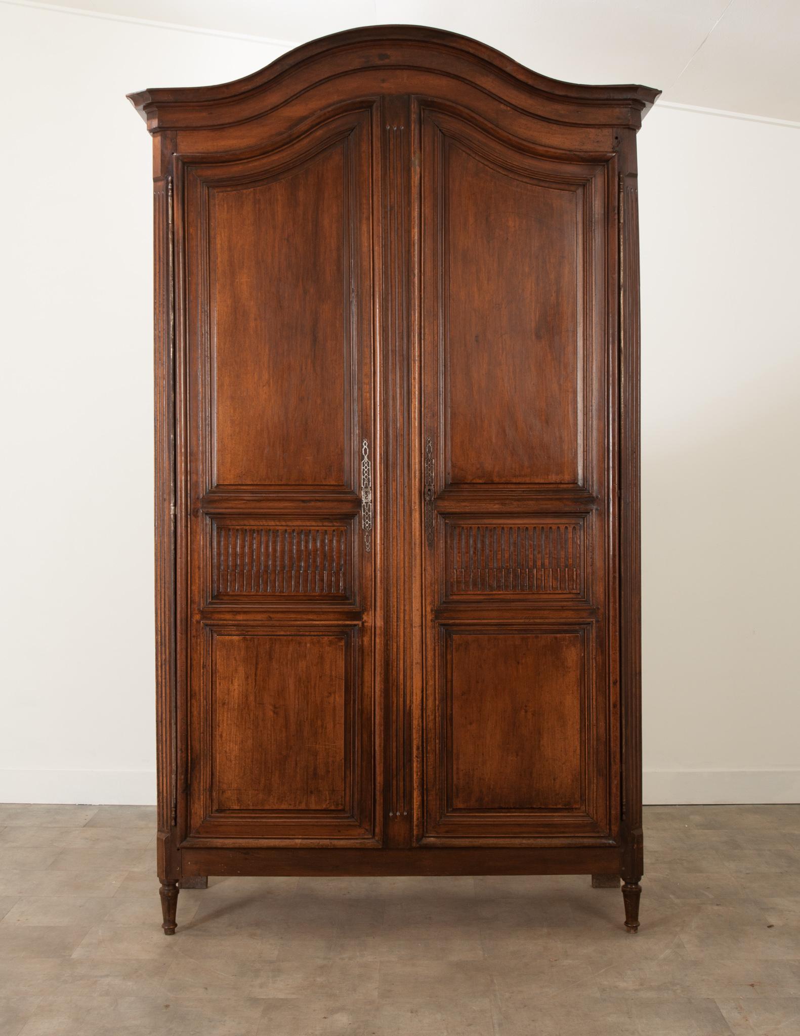 A massive solid walnut armoire from France, crafted during the 1840s. A chapeau de la champ cornice tops the armoire, heighting it to over 9 feet tall. The shapely paneled doors feature simple geometric carvings and hang on impressive steel barrel