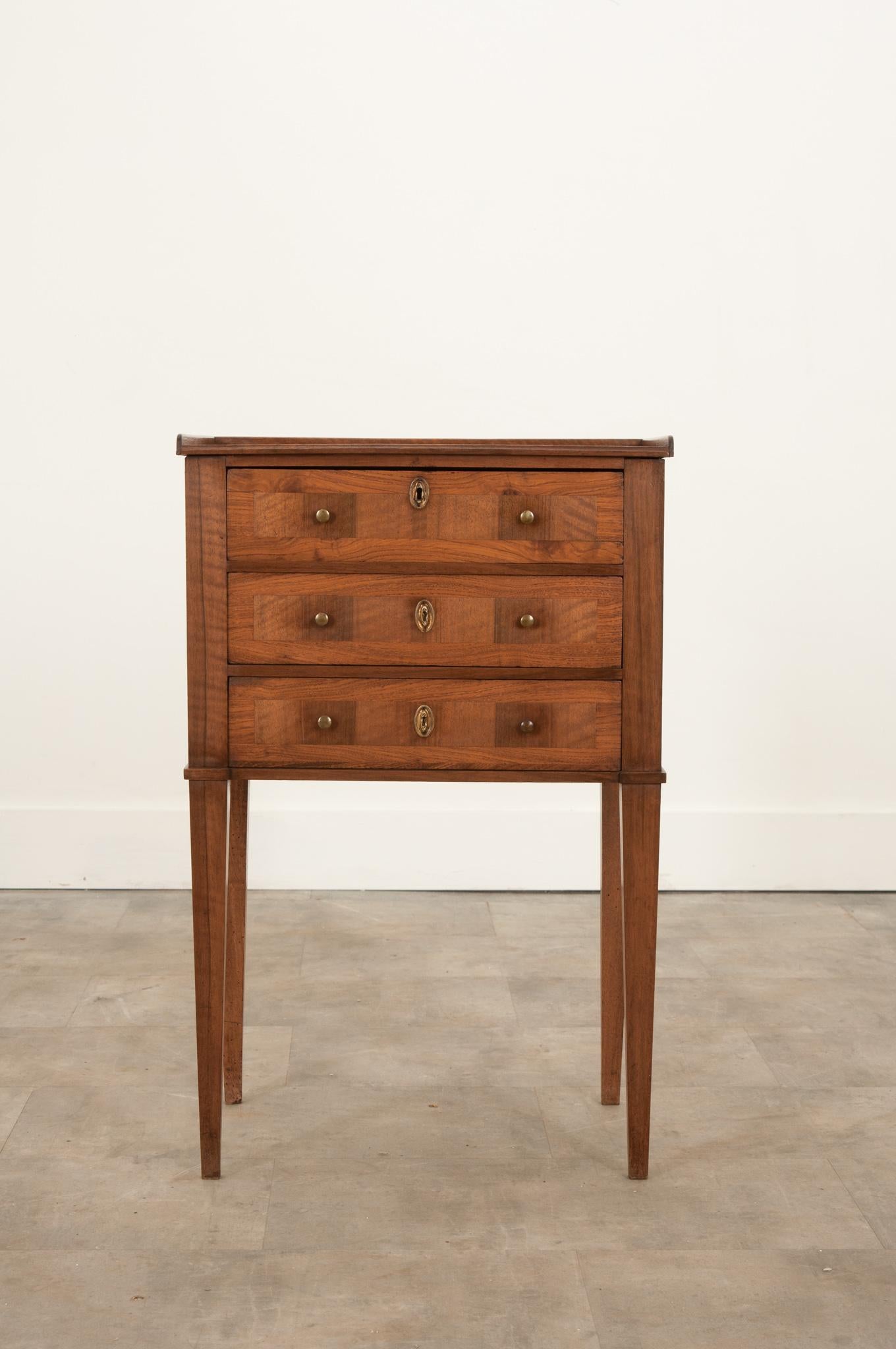 This darling petite commode was crafted in France, circa 1880, from a mix of vibrant woods. The top features a 3/4th gallery and molded edge. Three drawers each have a pair of simple brass pulls and an oval escutcheon. The top drawer neatly hides a