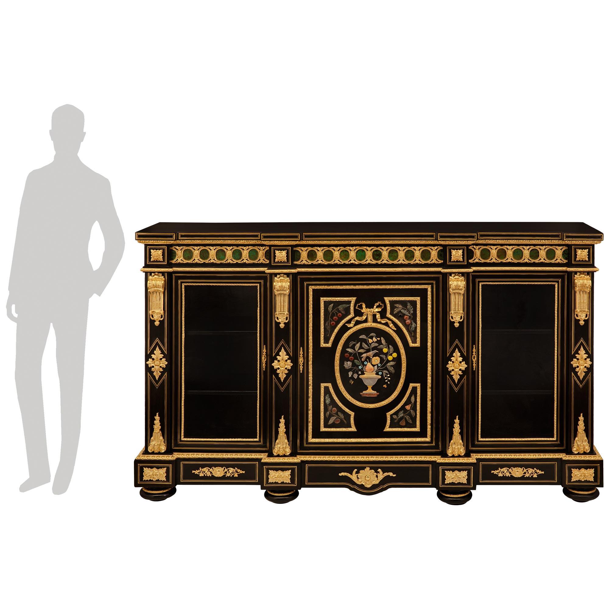 Spettacolare vetrinetta francese del XIX secolo del periodo Napoleone III in ebano, ormolu e pietra dura. Questo imponente mobile con intarsi Boulle è sollevato da sei piedini a baldacchino con fasce in Ormolu. Il fregio dritto e smerlato sopra