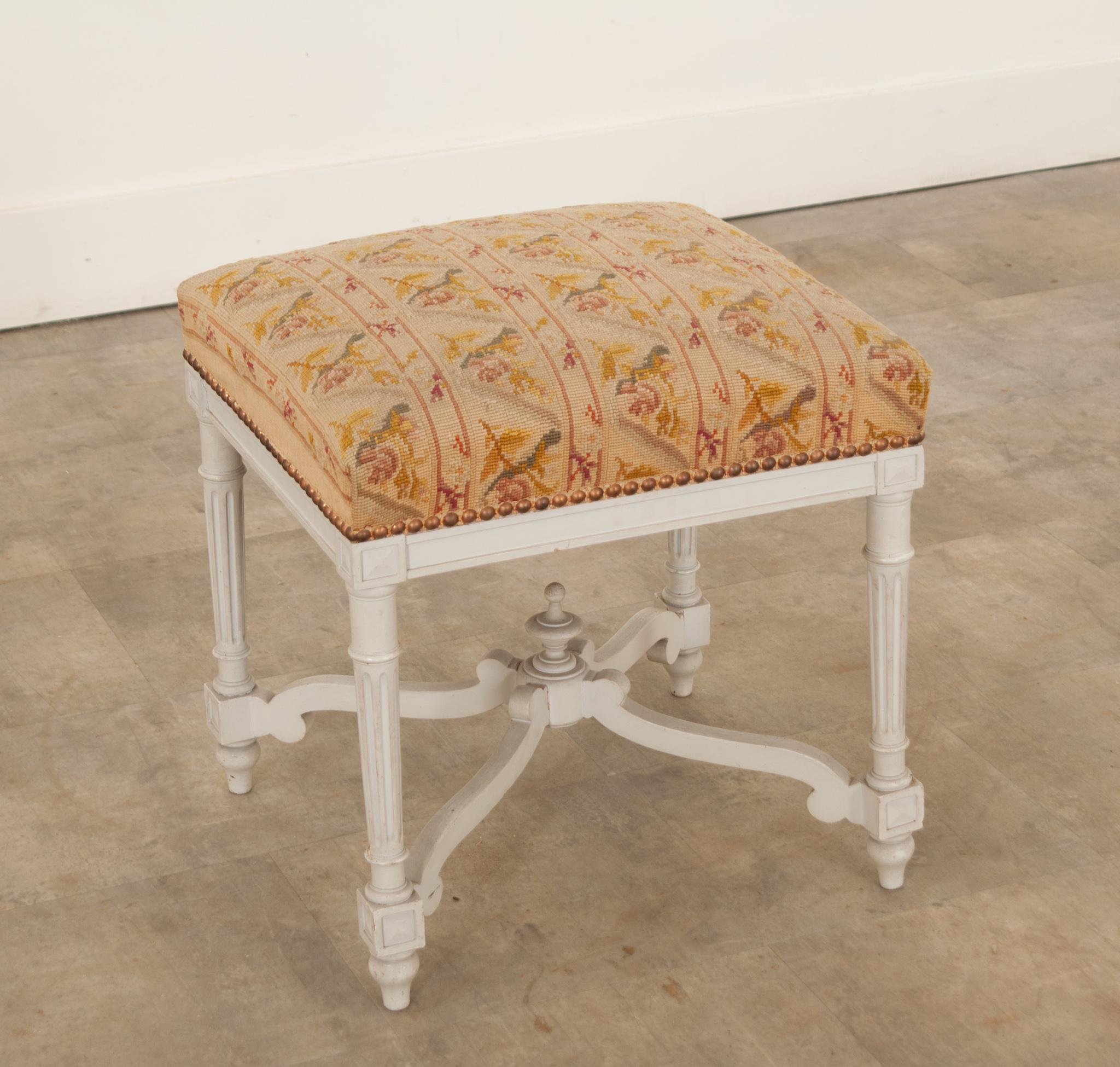 This delightful antique stool was hand-crafted in France, circa 1900. Square in shape, the stool stands on lovely turned and fluted legs connected to an elaborate, hand-carved, and shaped stretcher that meets in the center, topped with a decorative