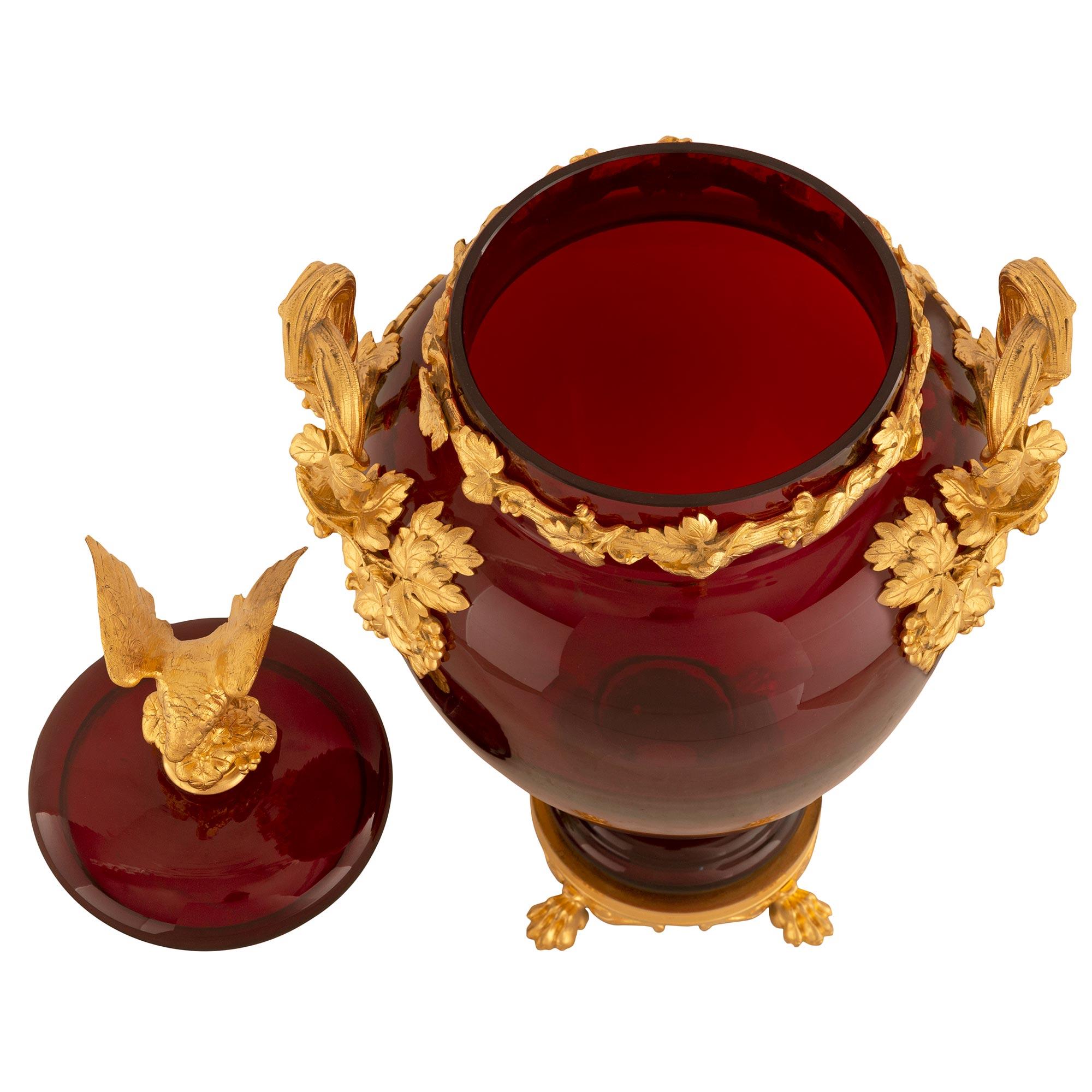 A stunning French 19th century Neo-Classical st. oxblood red glass and ormolu lidded urn. The urn is raised by a circular base with handsome ormolu paw feet amidst scrolled and fluted and hammered designs below the mottled socle shaped pedestal