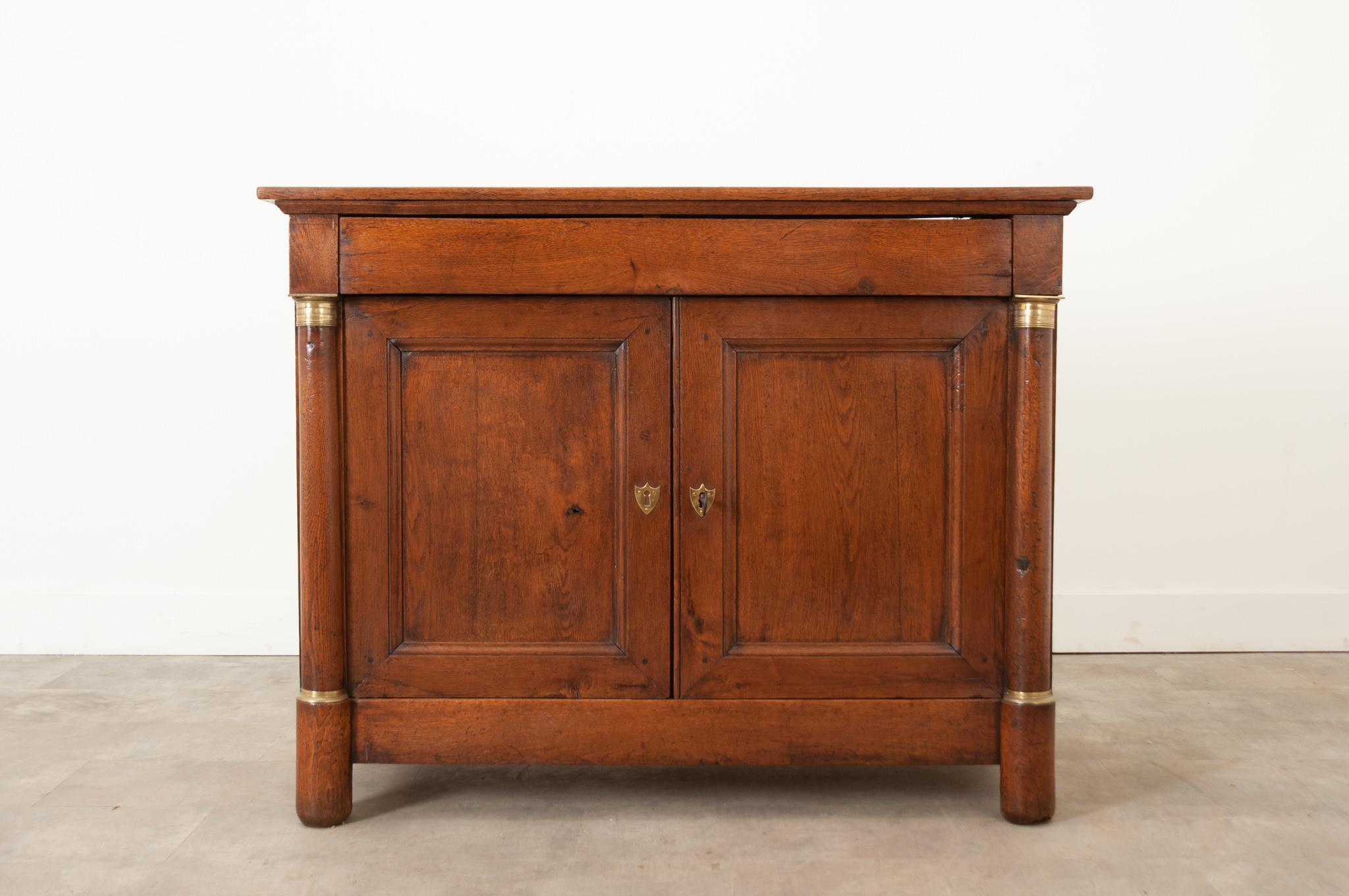 A more primitive Empire buffet made of solid oak is in wonderful antique condition. A long single drawer is located under the simple top. A pair of paneled doors are flanked by columns capped with gilt bronze ormolu at the top and bottom. Shield