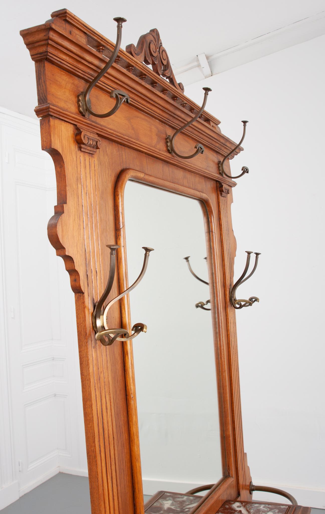 This handsome 19th century oak and brass hall tree is from France, circa 1880. The oak back is outfitted with three upper brass hooks and each side of the mirror is flanked with more for plenty of hat and coat storage. Mounted below is a brass rail