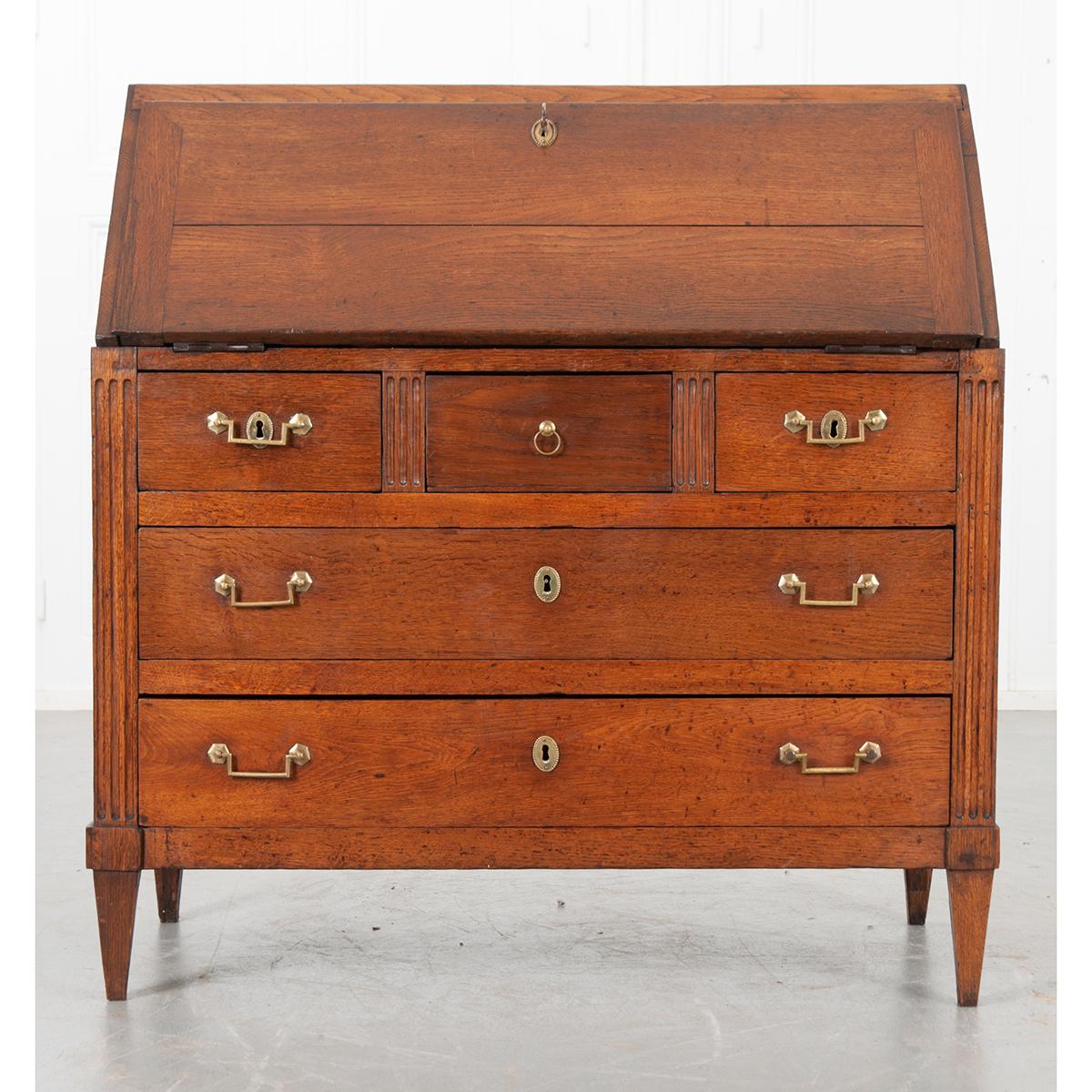This is a French 19th century Louis XVI-style desk made of oak. It has a slanted front that folds down to make a writing surface and reveals six small drawers with wood pulls and open storage areas. In the center compartment, the surface lifts to