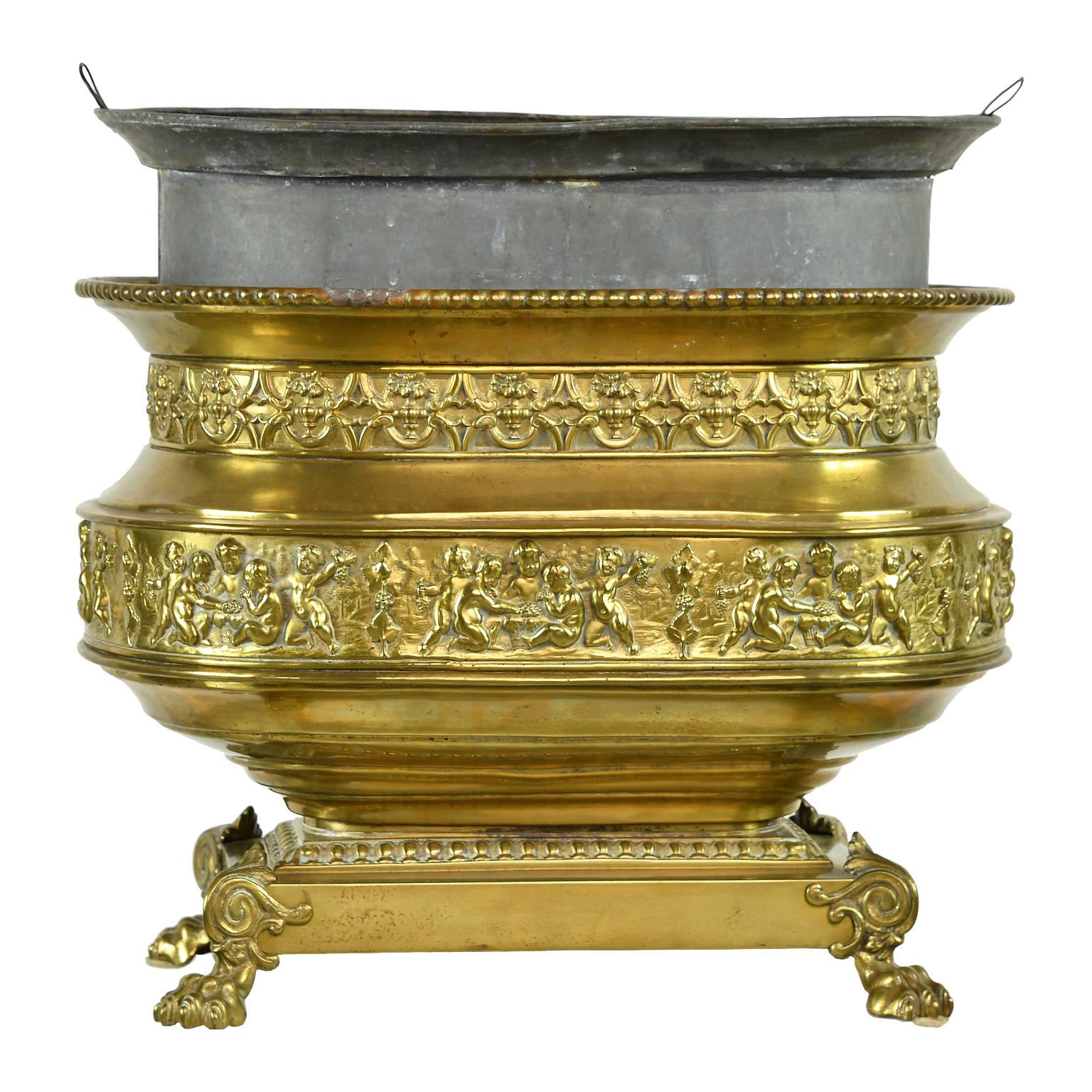 An exceptionally beautiful brass wine cooler or cache pot with oval urn resting on a rectangular pedestal base that is raised and supported by lions paw feet with wings. Features decorative bands of fine repoussé that include a Bacchanalian