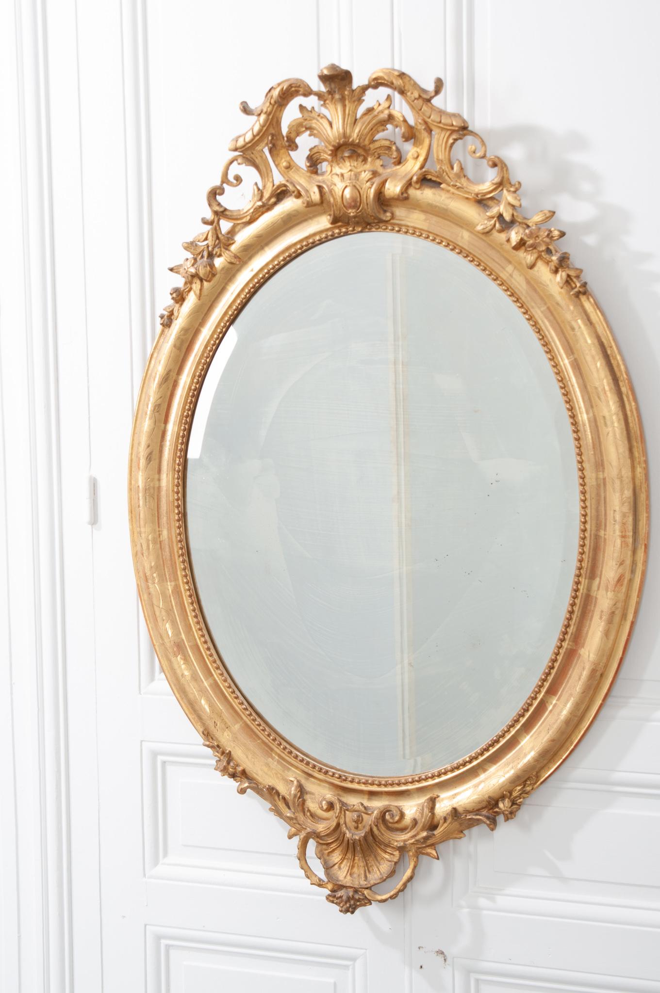 French 19th century oval gold gilt mirror with its original mirror plate. This mirror frame also has water gilting, adding another level of detail. It boasts intricate, carved details across the top and bottom, a beaded design around the inside
