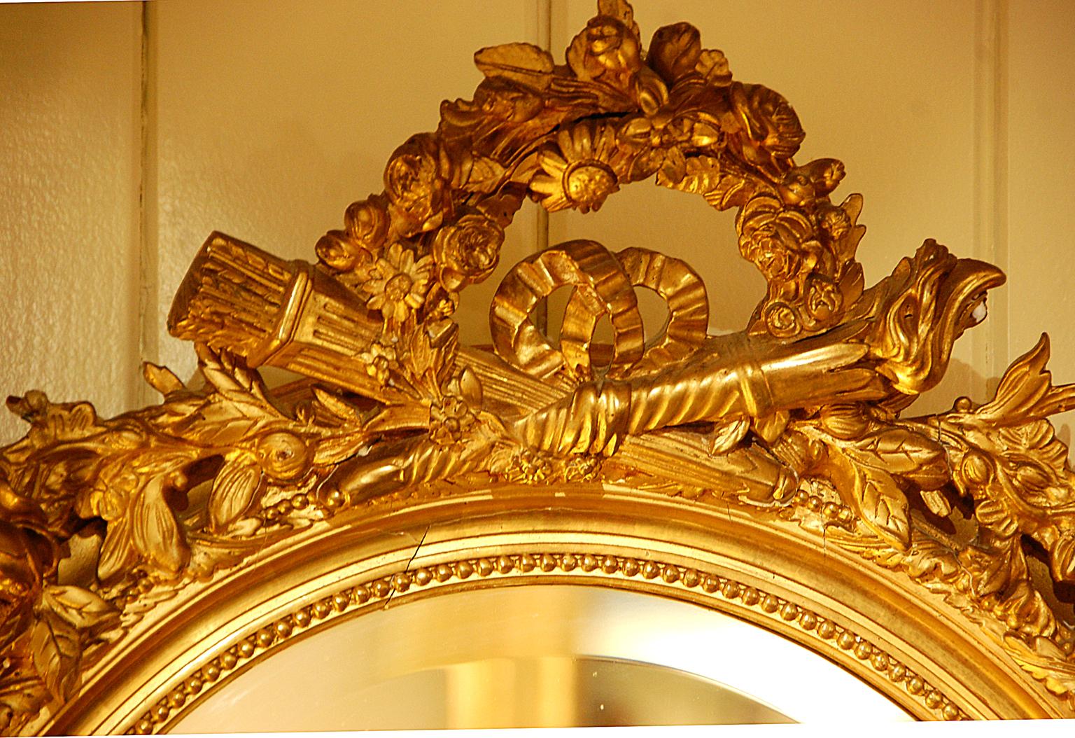 Miroir ovale français du XIXe siècle en feuille d'or vergée avec torche et carquois croisés entourés de motifs floraux dans le haut du miroir ; des feuilles et baies en volutes entourent le cadre et d'autres motifs floraux et feuillus dans le bas.