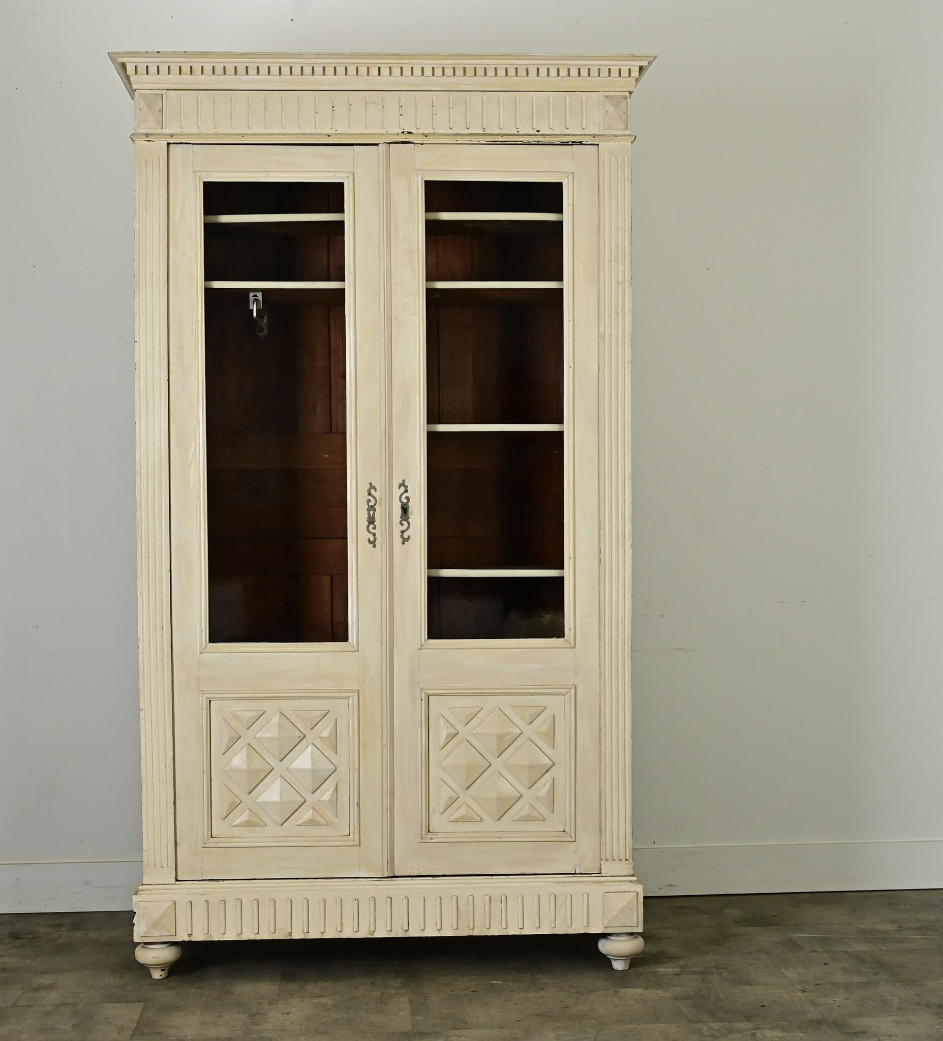 A French 19th century bibliotheque converted to an armoire with three-dimensional, geometric details and original paint in soft patina. Dentil molding in two scales lifts the narrow crown of the piece, enhancing the height along with fluted molding