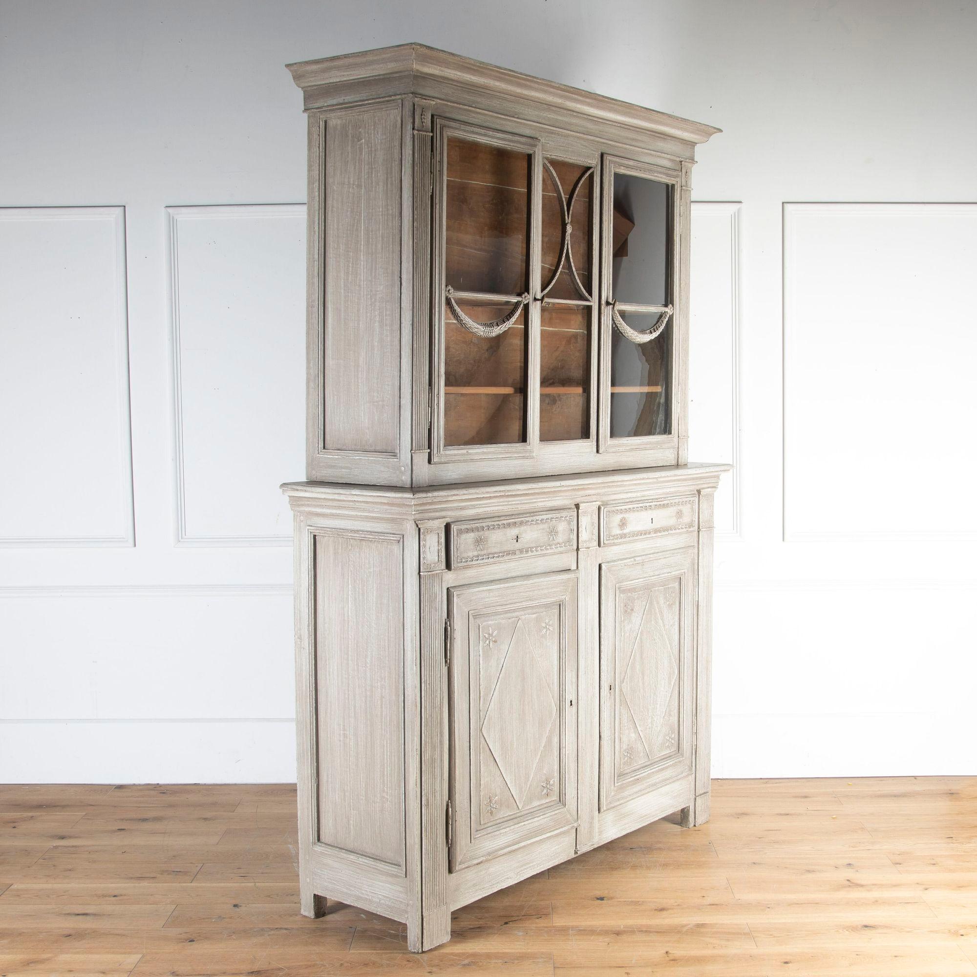 Early 19th Century French high-waisted cupboard.
This beautiful piece offers generous storage and unusual form. The top section with glazed doors featuring decorative carved swags and an unpainted interior offering several shelves. 
The bottom