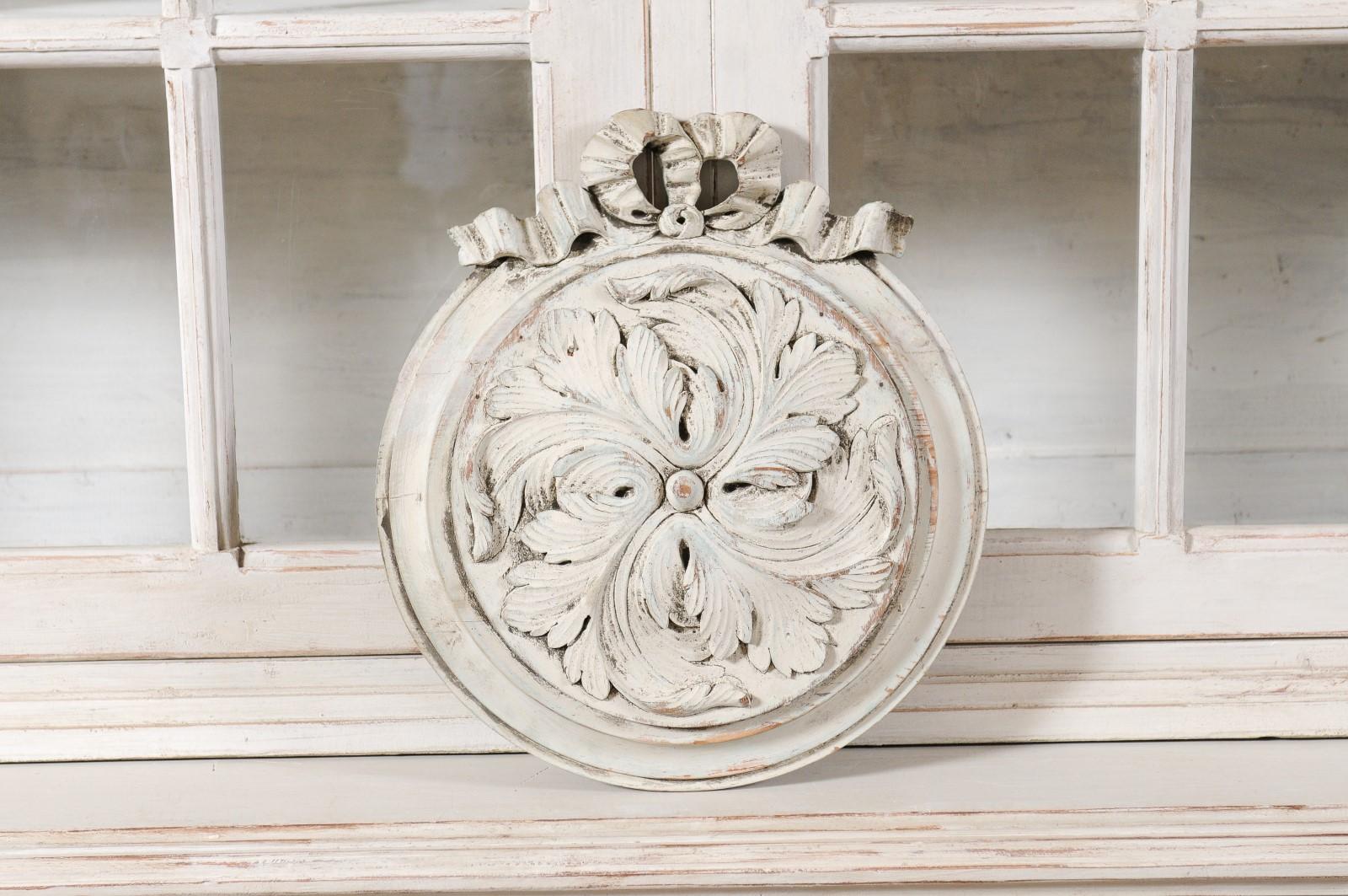 A French painted and carved wooden medallion from the 19th century, with low-relief acanthus leaves and ribbon motifs. Created in France during the 19th century, this lovely medallion features swirling acanthus leaves carved in low-relief, topped