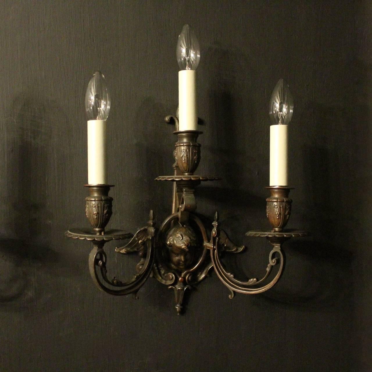 A French pair of cherub bronze triple arm antique wall sconces, the leaf square gauge scrolling arms with circular reeded bobeche drip pans and decorative floral bulbous candle sconces, issuing from winged shield backplate with a lovely central