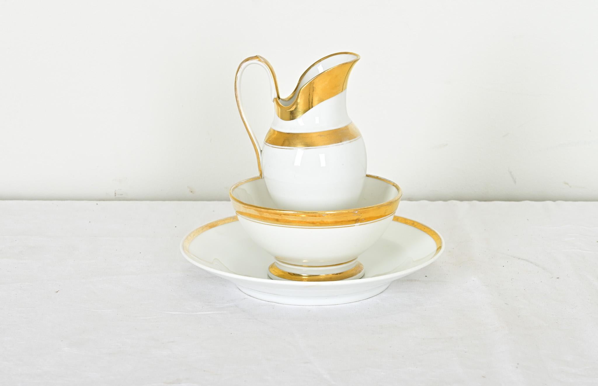 An elegant, three-piece set of old Paris porcelain in the style of Lebon-Halley with a pitcher, bowl, and deep plate in creamy porcelain accented by luxurious gold trim. The underside of the pitcher is marked with a “49” in the bottom and a “10”