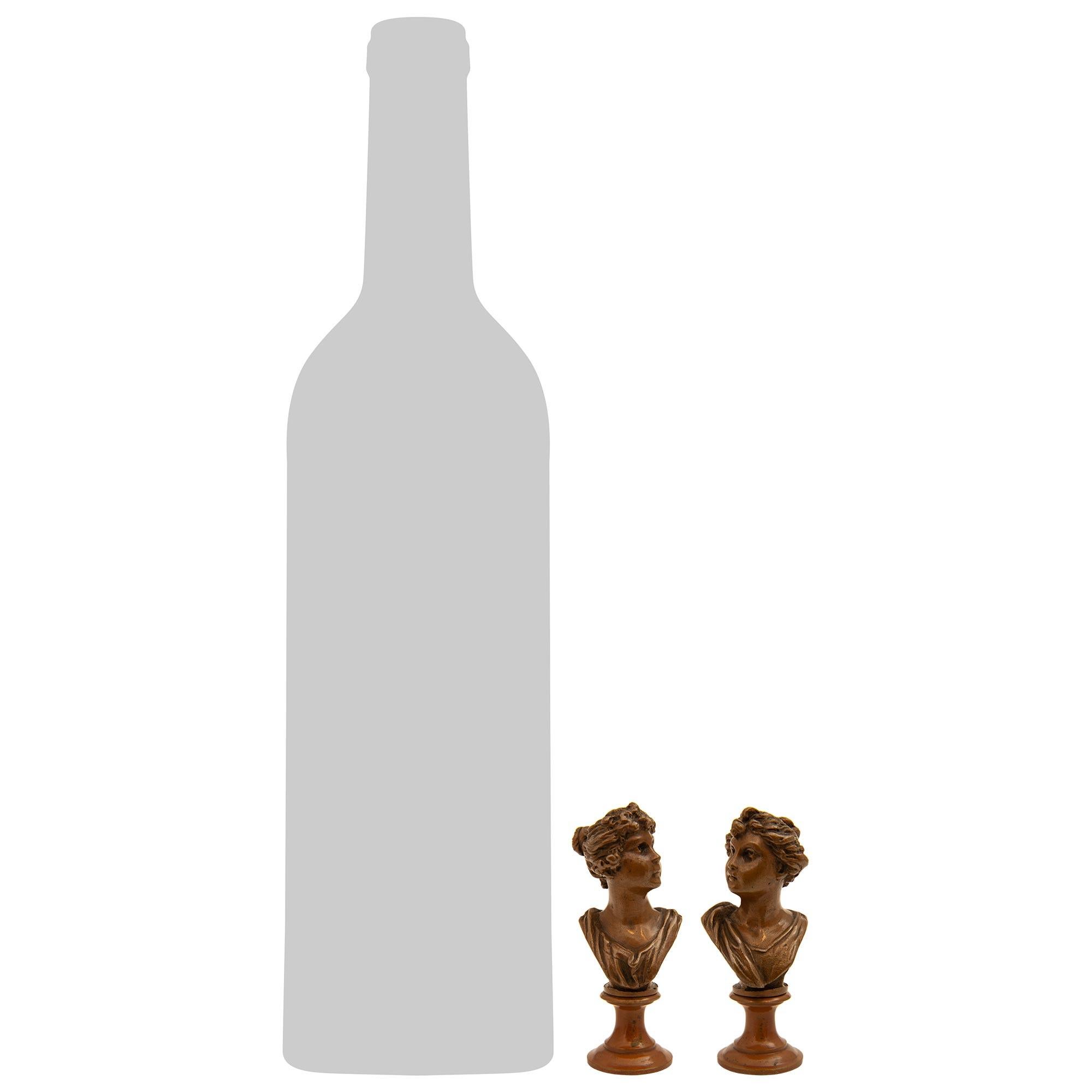 Una bella e piccola coppia di busti in bronzo patinato francese del XIX secolo. Ciascun busto in bronzo è innalzato su un piedistallo circolare a socle screziate. Le eleganti fanciulle qui sopra sono vestite con abiti classici e fluenti, con i