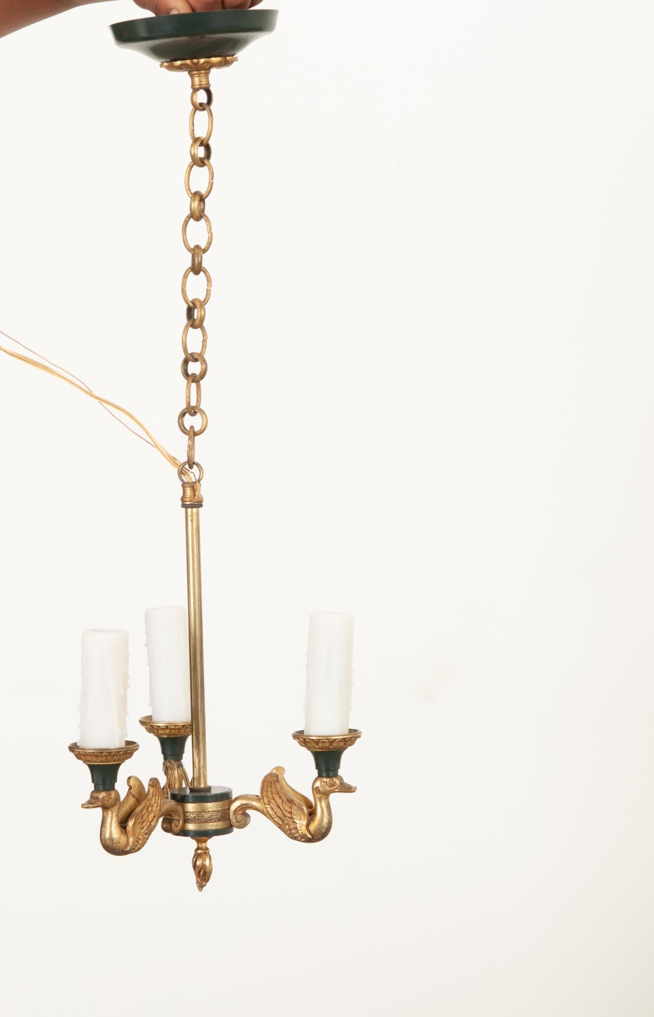 This darling petite Empire style chandelier is just the fixture your space needs to elevate it to the next level. Cast brass swans support three arms and attach seamlessly to a decorative brass and bronze center ring. White, faux melting candle