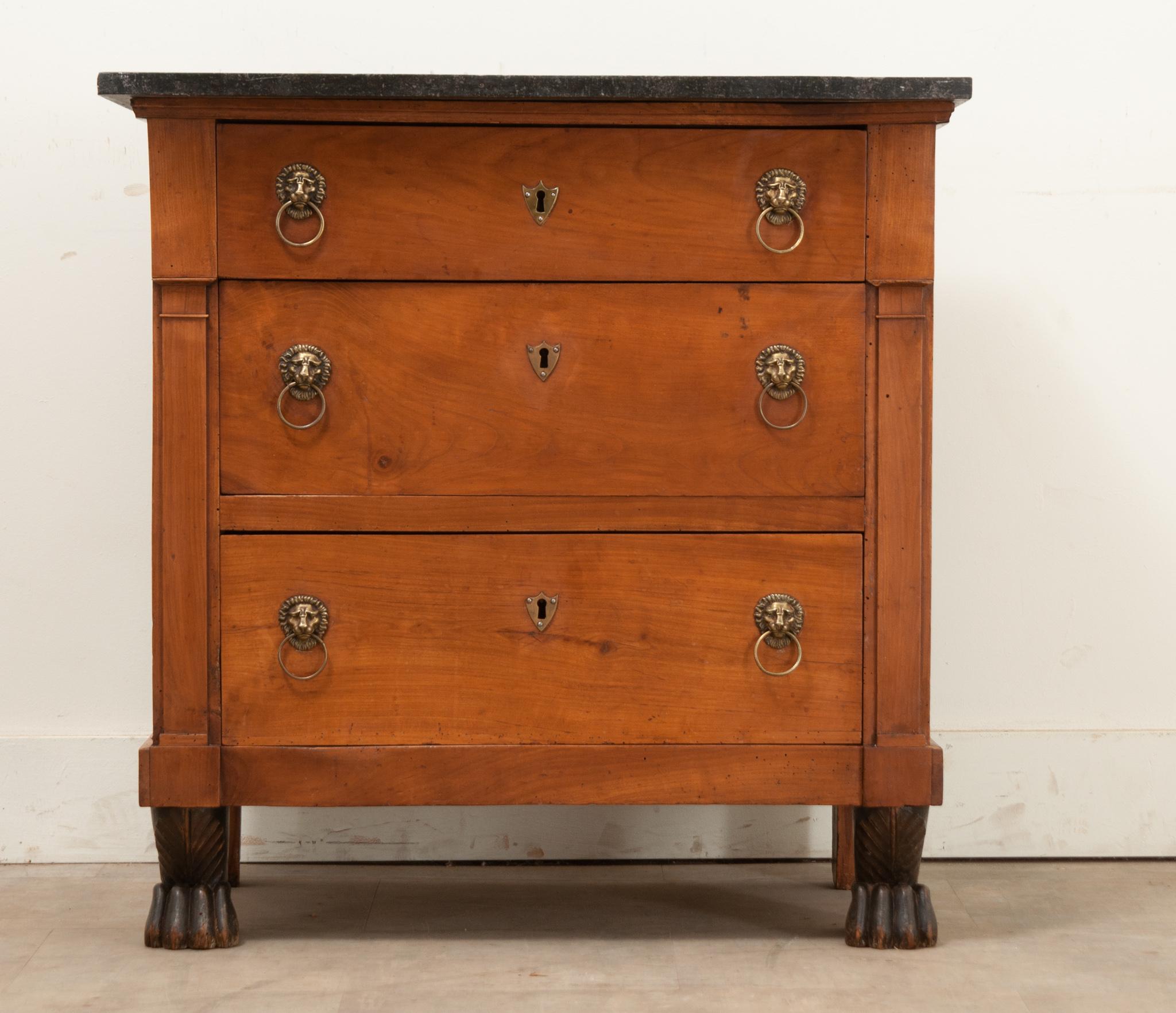 A petite French Empire commode made of mahogany, is topped with its original black fossil marble. Three easy-to-use drawers with brass lion head pulls are flanked by column form legs ending in ebonized paw feet. Cleaned and polished with a French