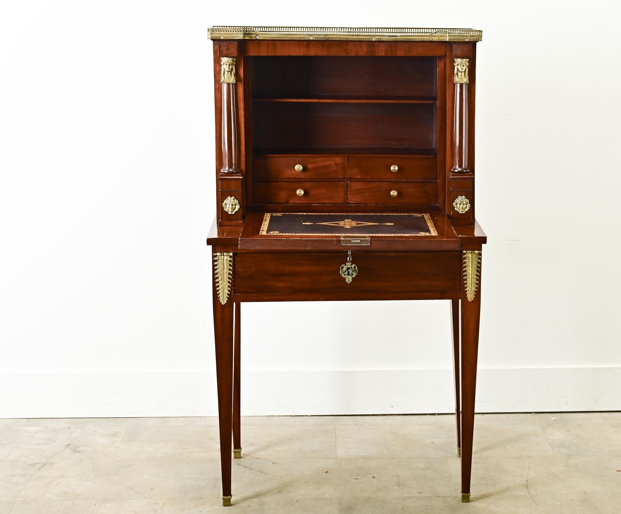 A petite French Empire mahogany secretary or lady’s correspondence desk. This secretary has an inset black fossil marble top surrounded by a pierced brass gallery. Below is a mirrored fold front door that opens with a working lock and becomes the