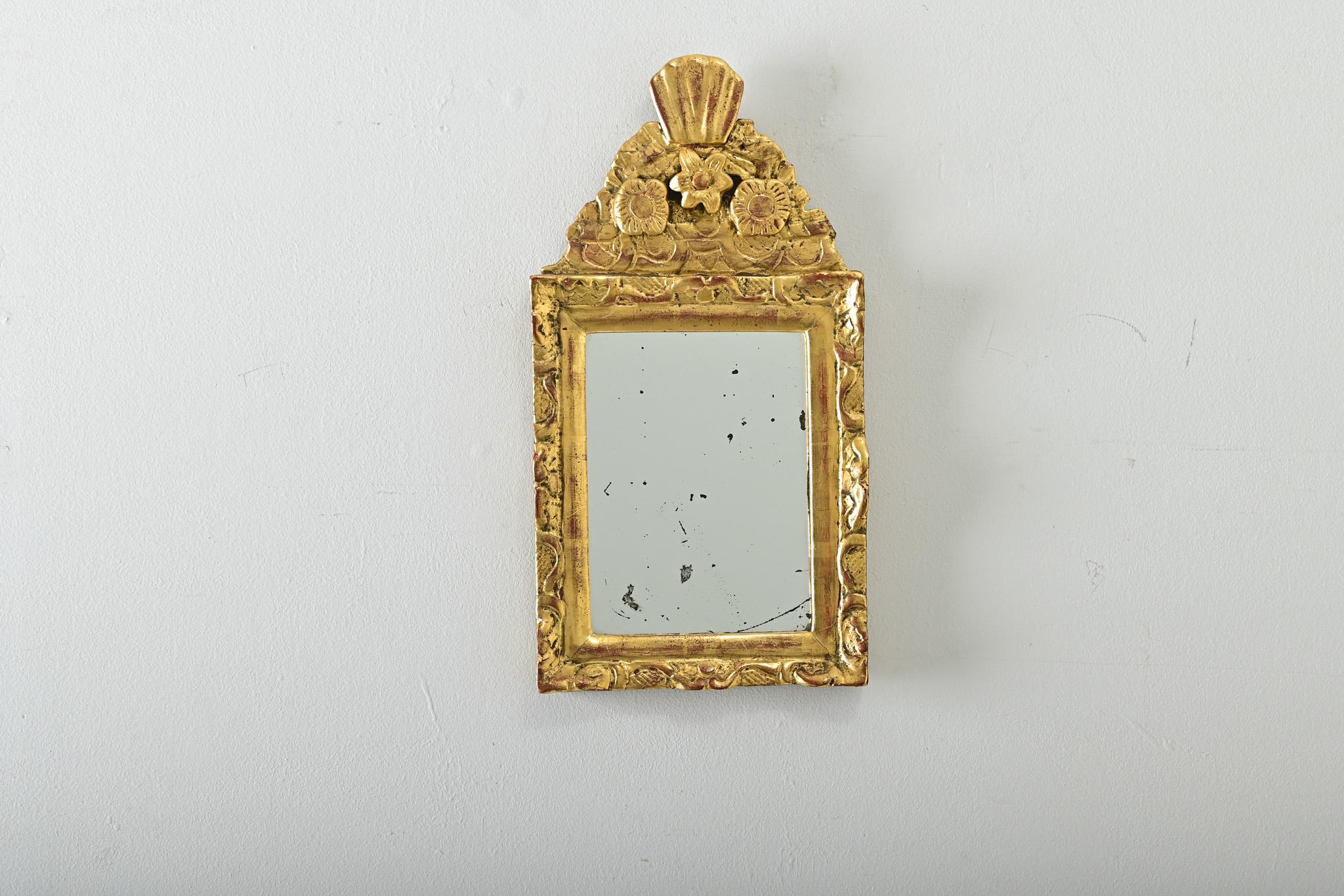 Pequeño espejo francés de estilo Luis XVI, de hacia 1870, con lustrosos detalles de madera dorada tallada que irradian un efecto mucho mayor que sus diminutas proporciones. En la parte superior de la pieza, una corona festoneada se eleva sobre un