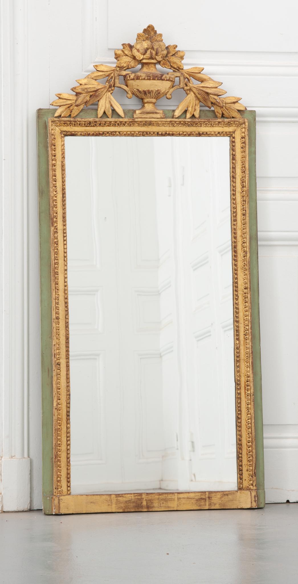 A lovely petite 19th century French mirror, made in the style of Louis XVI. Circa 1840, the worn gold gilt and sage green paint have gained a wonderful patina. A beautiful pierced carving of an urn overflowing with flowers and laurel sits atop the
